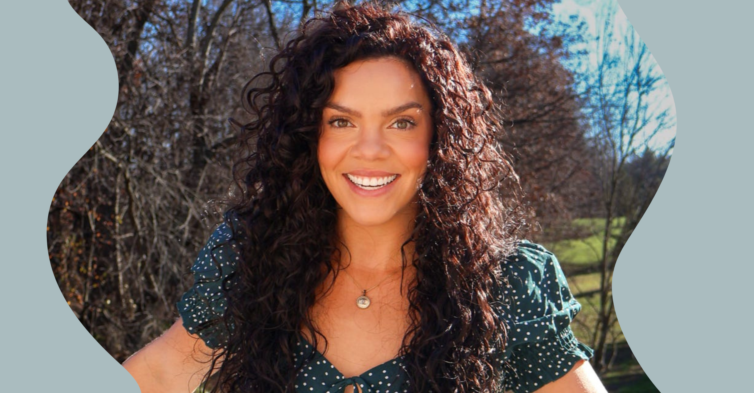 Close-up of a woman with curly dark brown hair smiling outdoors on a sunny day, wearing a dark blue polka dot blouse and a necklace, with a background of leafless trees and blue sky.