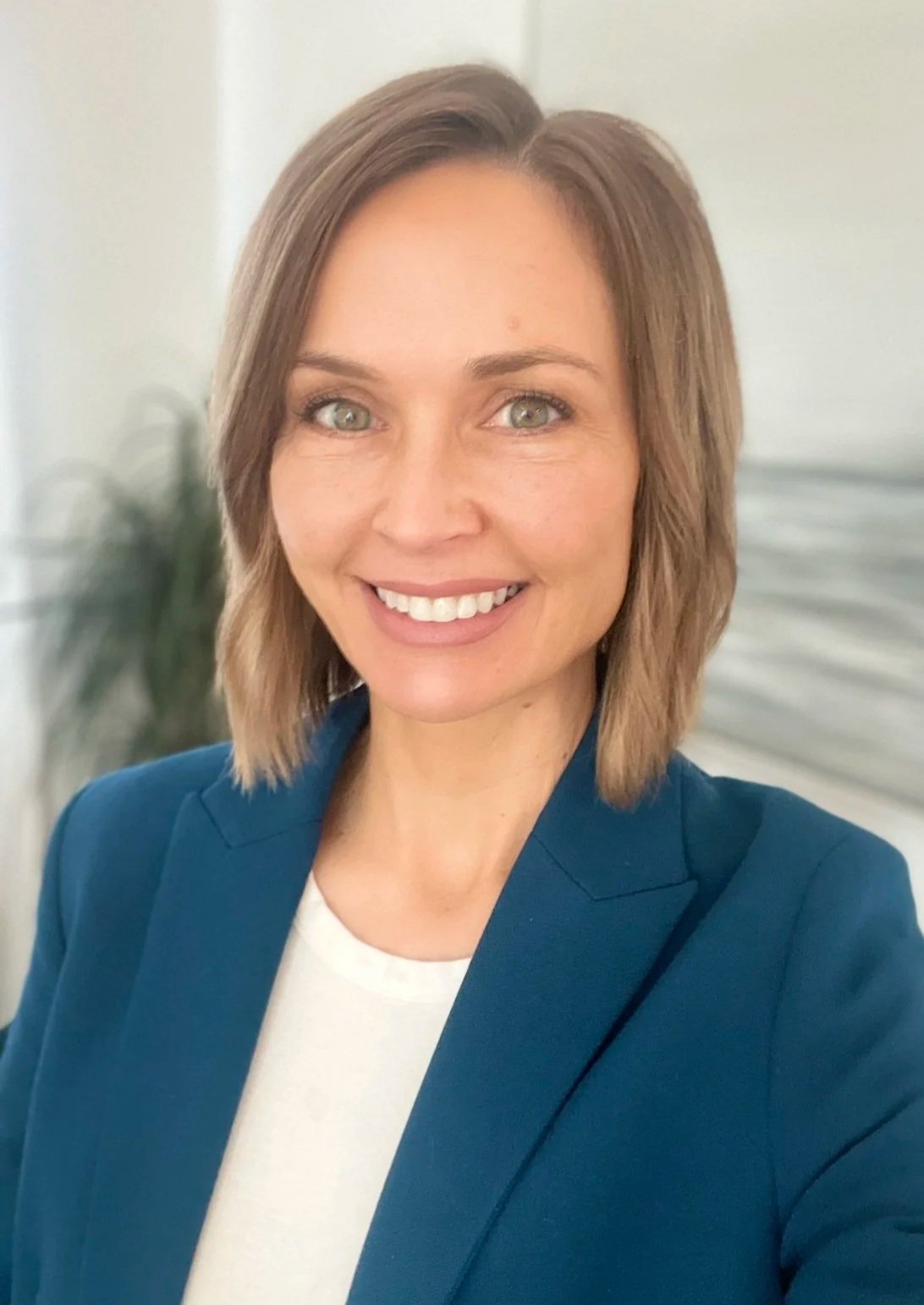 A woman with shoulder-length brown hair and blue eyes taking a selfie indoors, wearing a blue blazer over a white top, smiling at the camera with a blurred background of a houseplant and white wall.