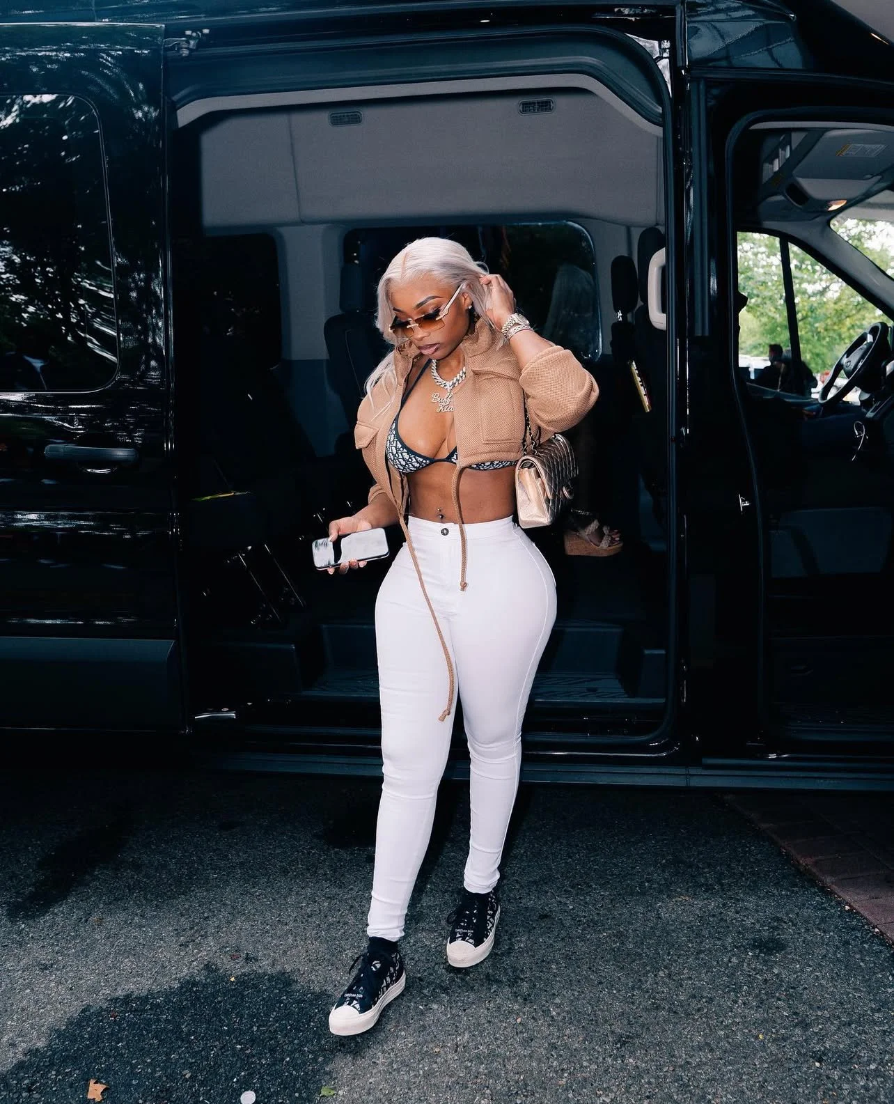 A woman with platinum blonde hair wearing sunglasses, a bra top, white pants, and sneakers standing outside a black van, looking at her phone.