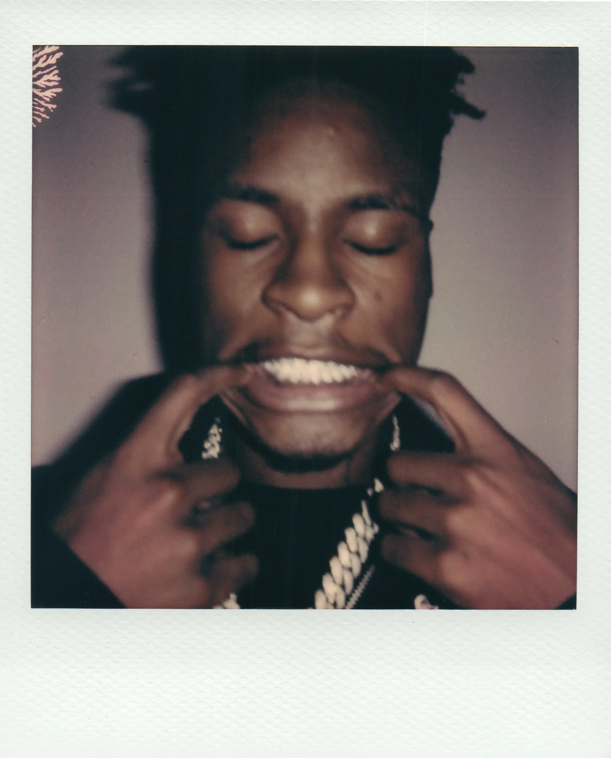 A young person with a short afro hairstyle pulling a big smile, showing their teeth, while using their fingers to stretch their mouth wider. They are wearing a black shirt and a chunky chain necklace.