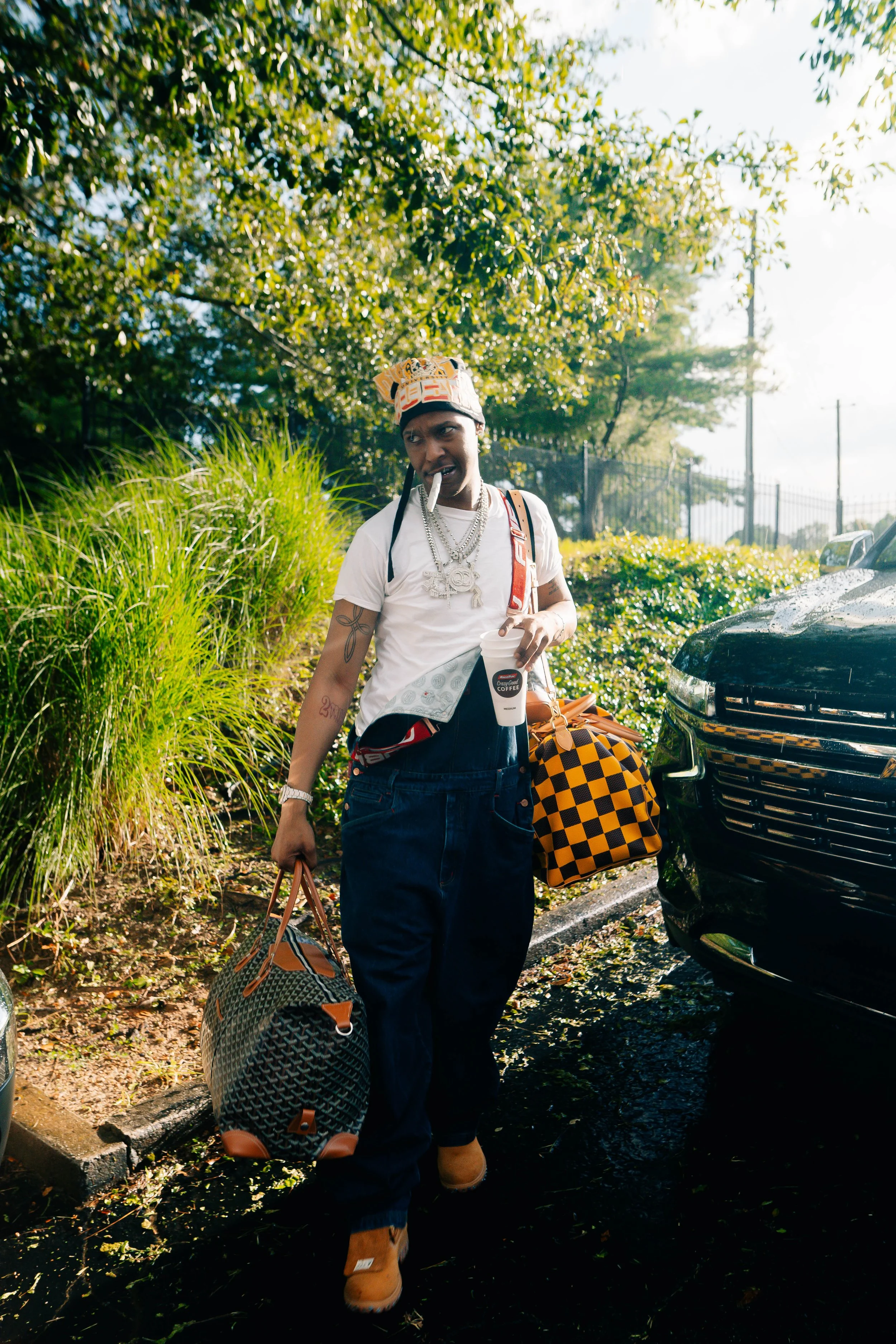 A young man with tattoos, wearing a white T-shirt, dark baggy jeans, and tan shoes, holding a large travel bag and a small checkered bag, walking outdoors near a car on a sunny day, with trees and greenery in the background.