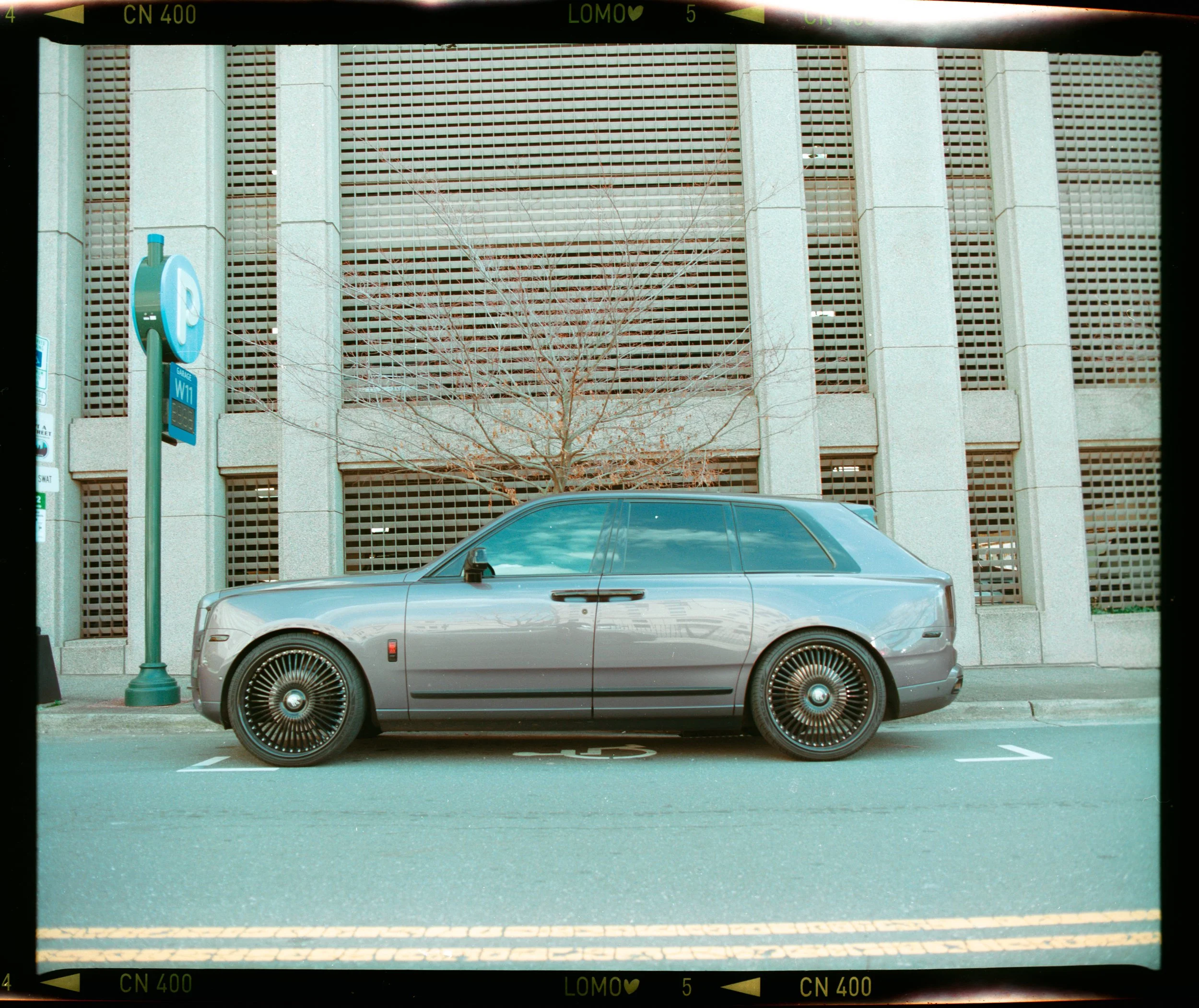 Silver luxury sedan parked in a parking space near a parking meter and an urban building with concrete pillars and lattice design. A leafless tree is visible behind the car.