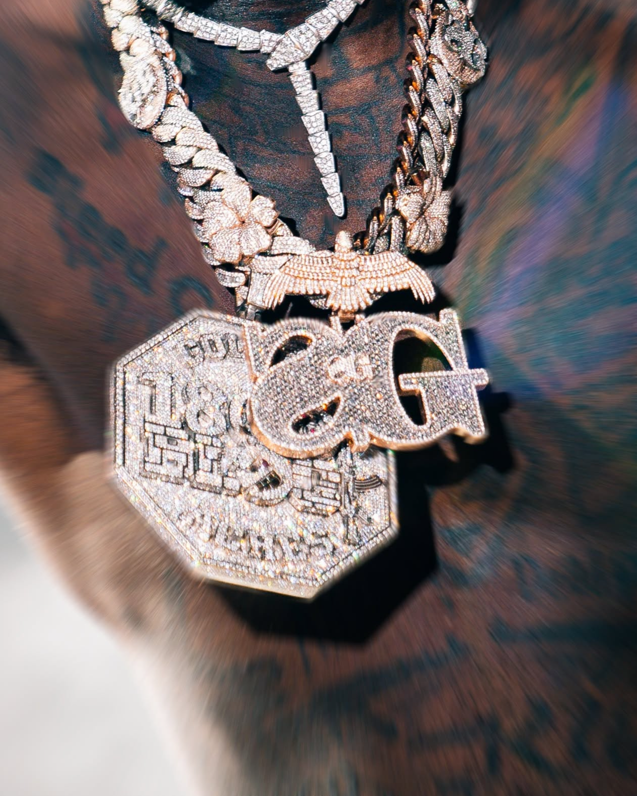 Close-up of diamond-encrusted jewelry, including a large pendant with the word 'GOD' and an eagle, worn over a dark, sheer fabric.