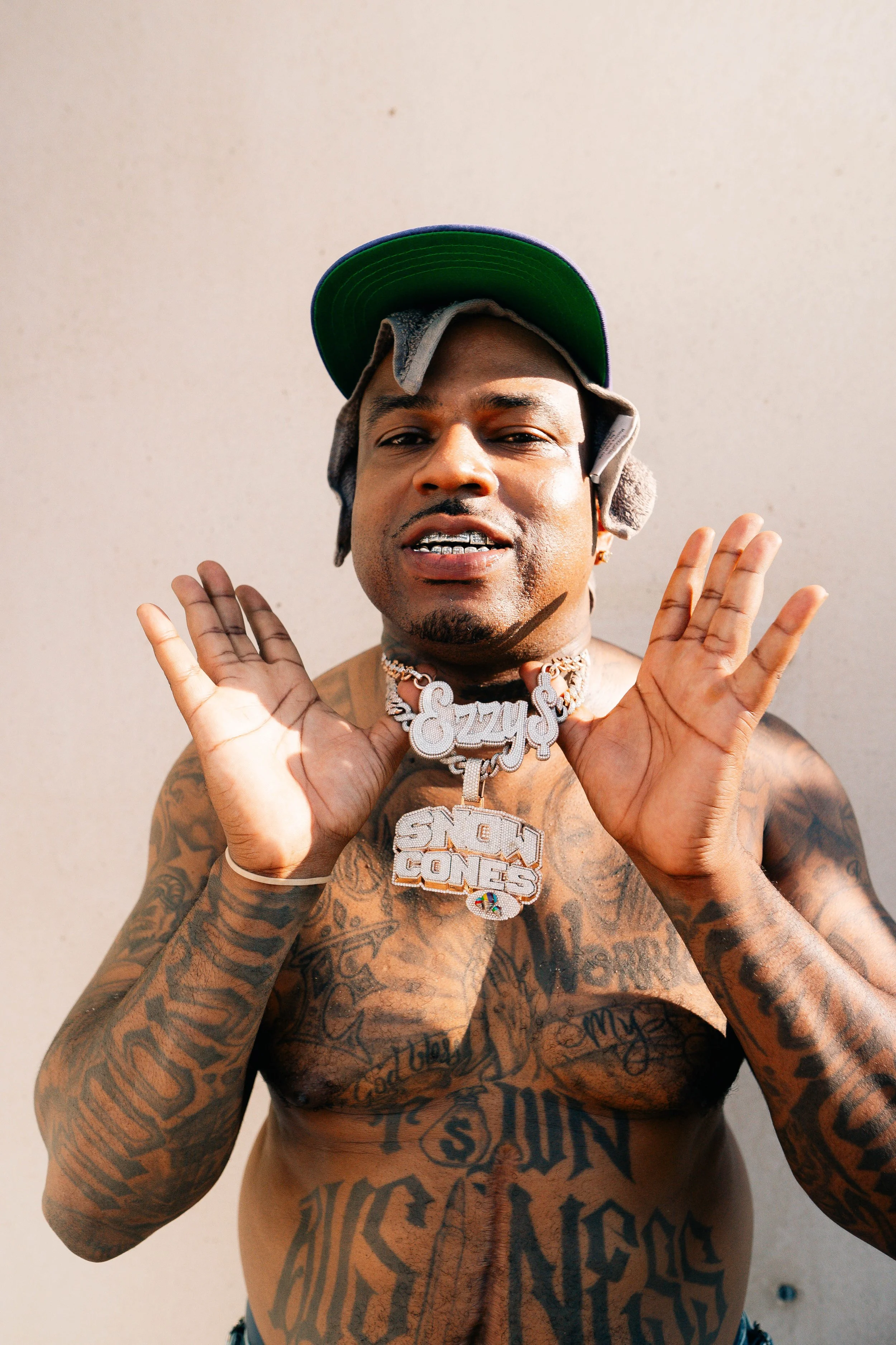 A shirtless man with tattoos, wearing a green cap and large, jewelry necklaces, standing against a plain background.
