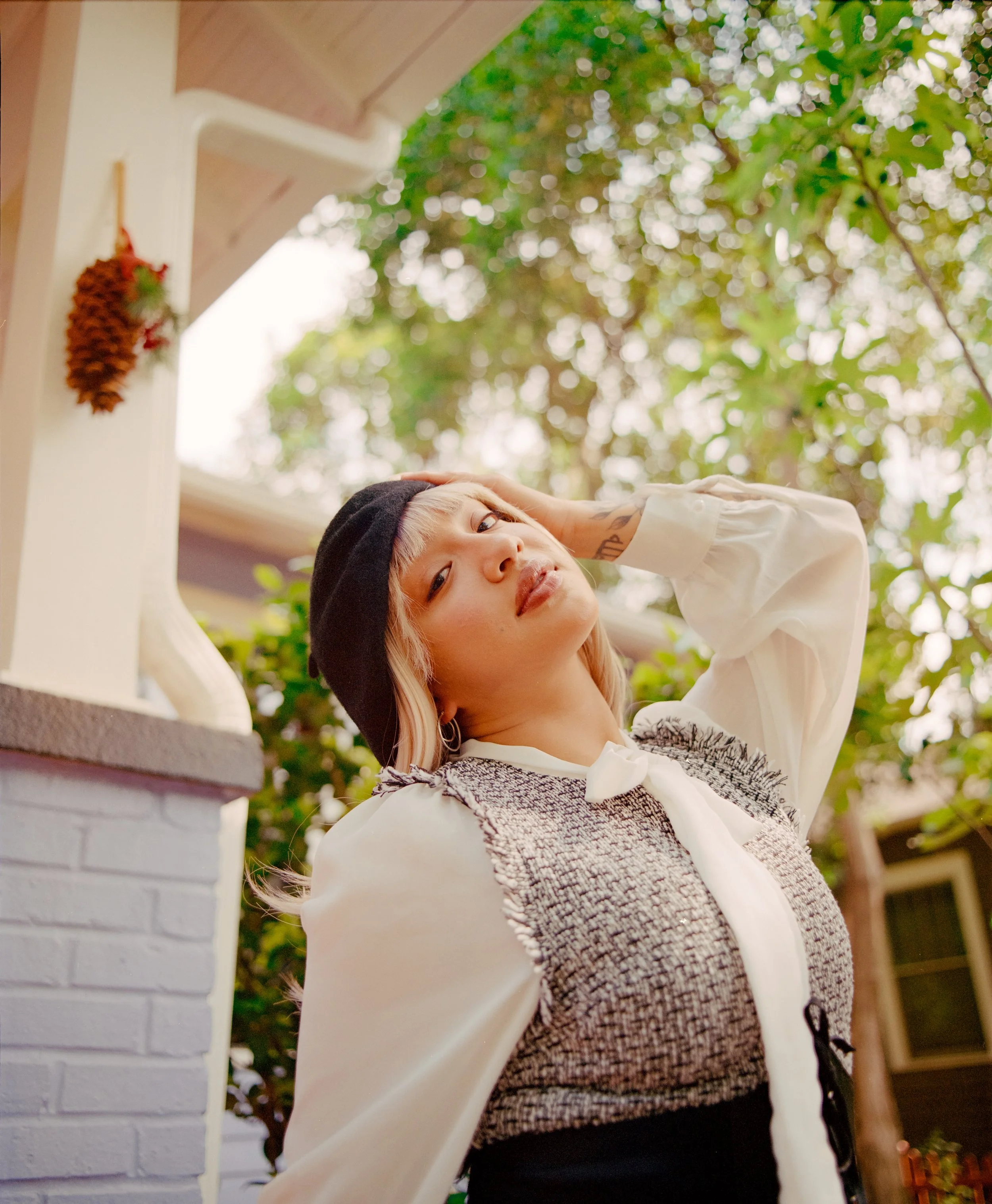 A woman with blonde hair and a black beanie posing outdoors, with greenery and part of a house visible in the background.