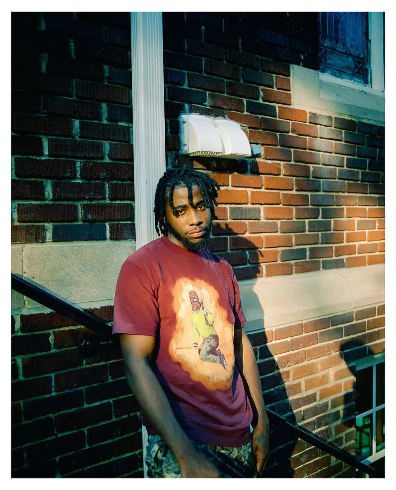 Young man with dreadlocks leaning against a black railing outside, wearing a maroon T-shirt with a graphic of a character holding a microphone, set against a brick wall with a window and white utility box.
