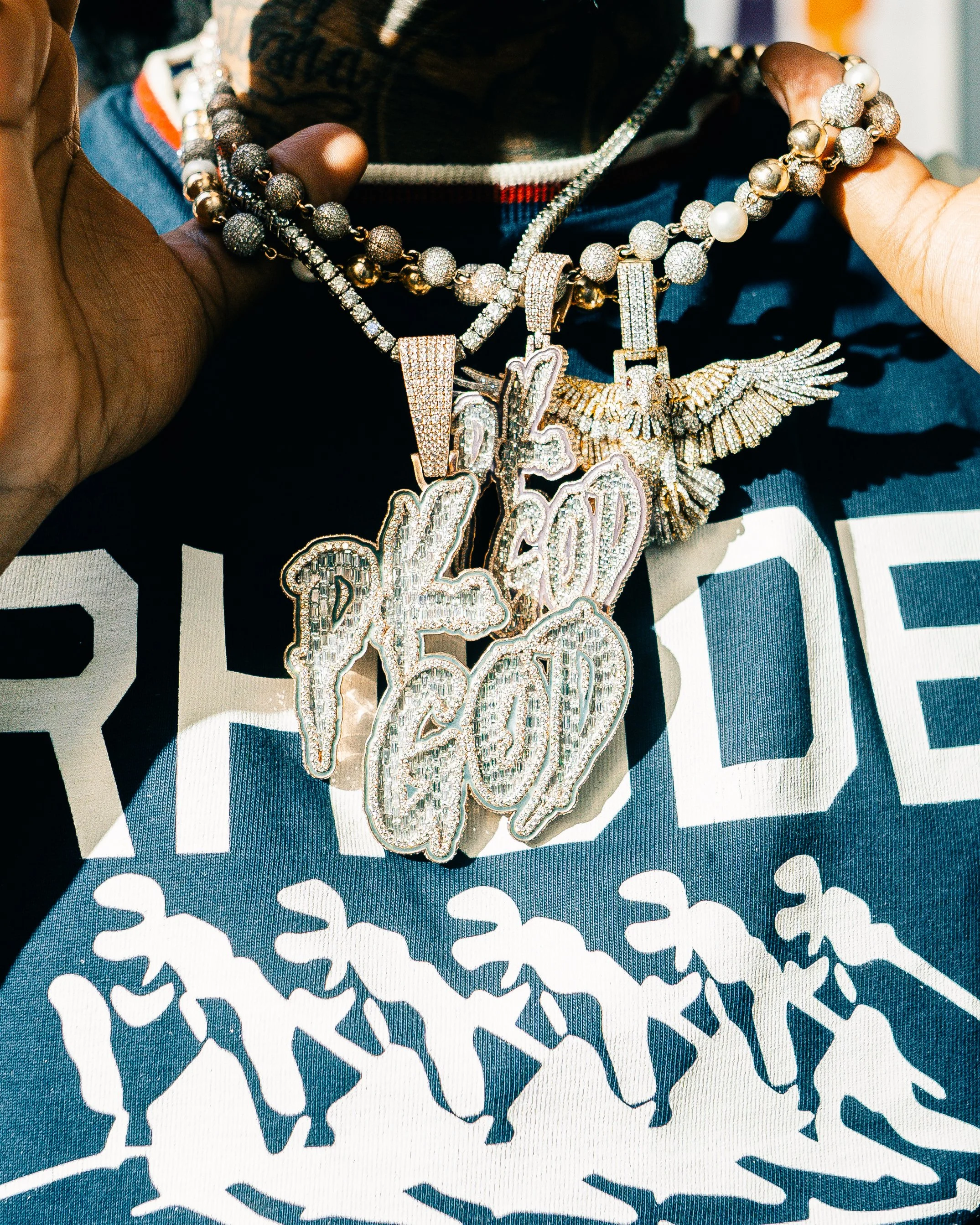 Close-up of a person wearing multiple luxury necklaces with large, jeweled pendants, including words and symbols, over a printed shirt.