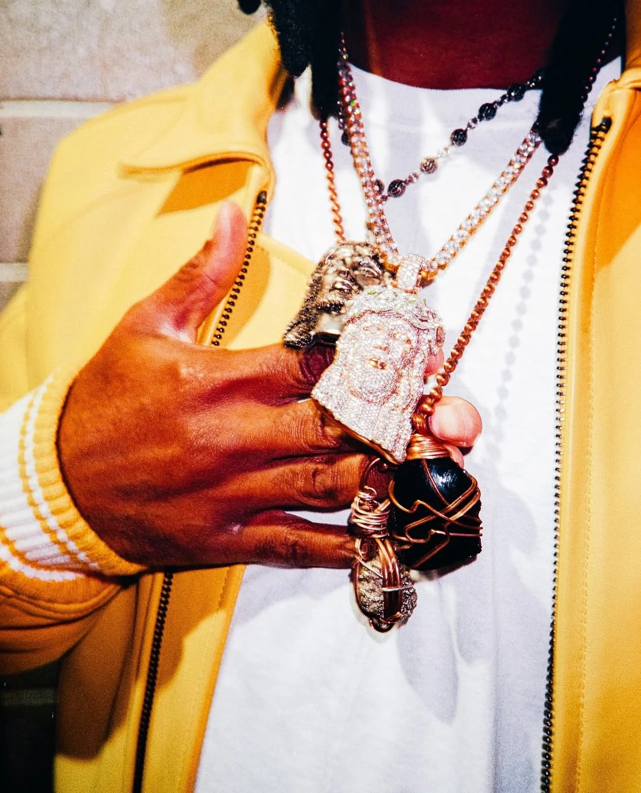 Person wearing a yellow jacket and white shirt holding multiple gold and diamond chains with large pendants, including a face and a lion's head.