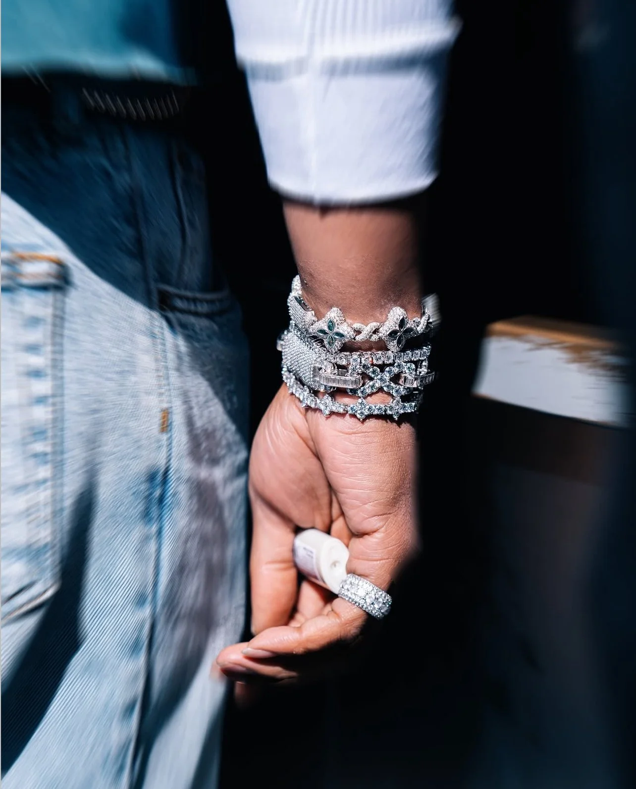 Close-up of a person's hand adorned with multiple silver bracelets and a large diamond ring, holding a white object, with the person wearing a white and blue striped shirt and light blue jeans.