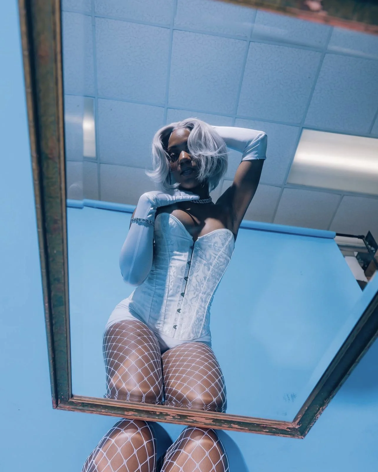 A woman with platinum blonde hair in a wavy bob, wearing a white corset, white gloves, fishnet stockings, and accessories, poses in front of a mirror ceiling with a blue wall background.