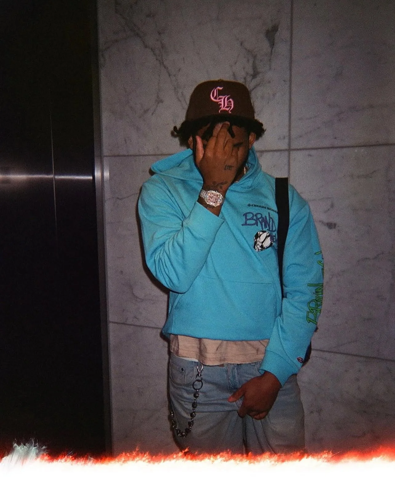 A man wearing a baseball cap, blue hoodie, and jeans standing against a marble wall, covering part of his face with his hand.