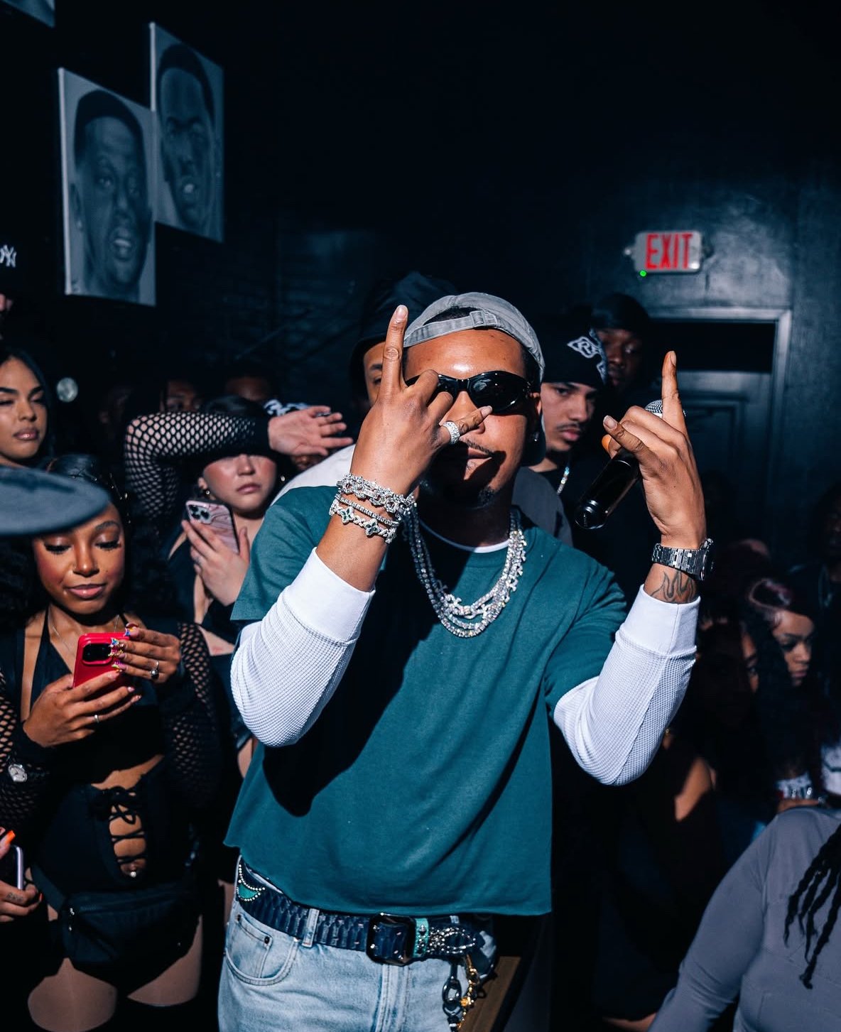 A man wearing sunglasses, a gray cap, jewelry, and a multicolored shirt making a hand gesture at a crowded party, with people taking photos and a dark wall decorated with black and white portraits in the background.