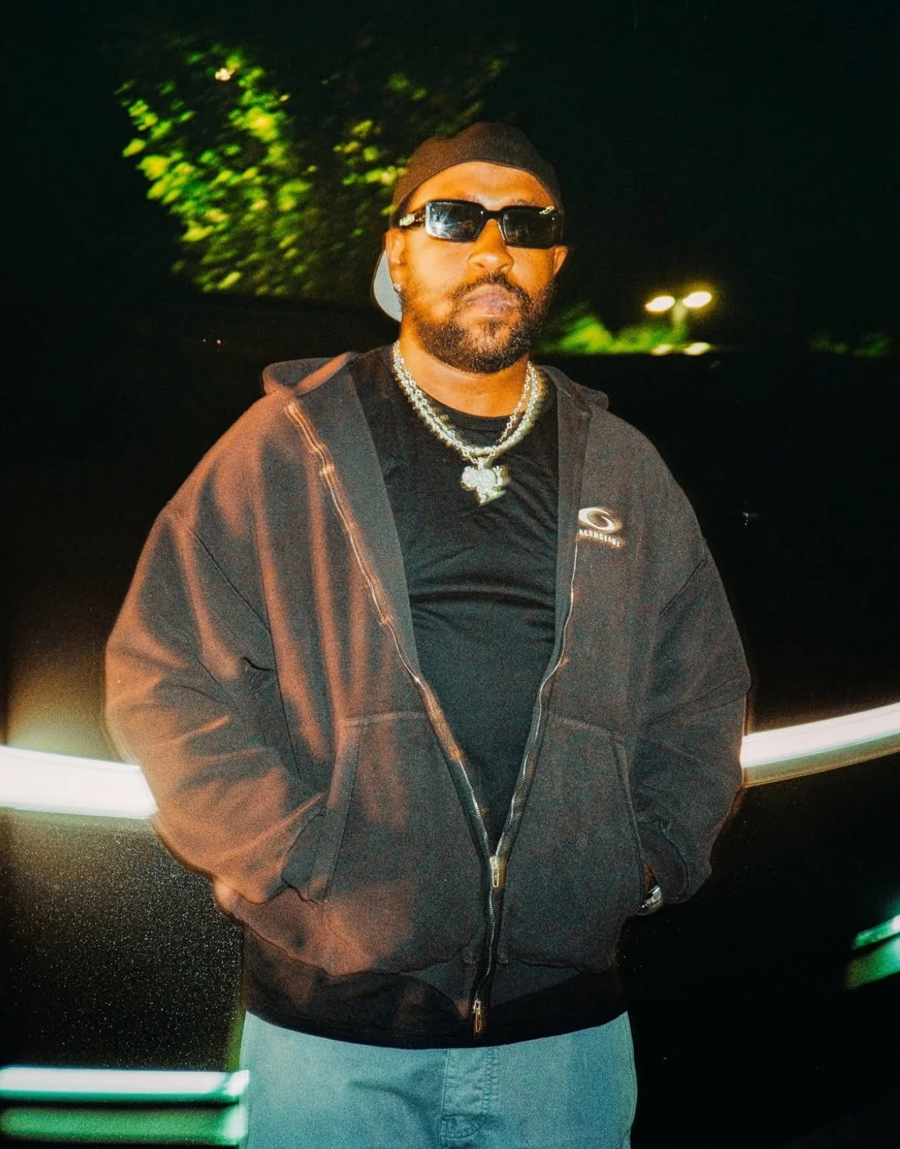 A man with sunglasses, a beanie, and a black hoodie stands outdoors at night with his hands in his pockets