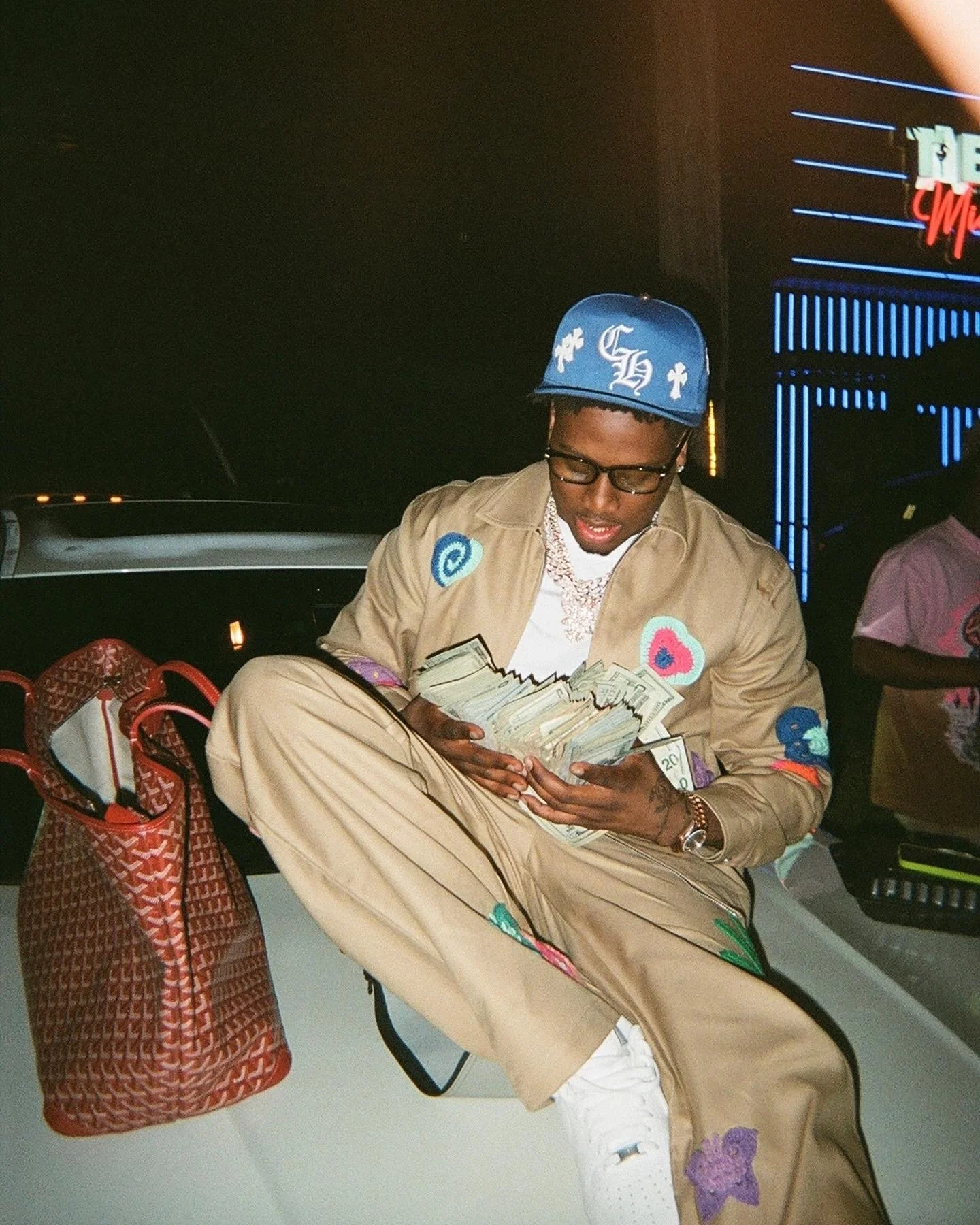 A young man sitting on the hood of a car at night, counting a large stack of cash, wearing a tan outfit with colorful patches, a blue baseball cap, sunglasses, and jewelry, with a purse and neon signs in the background.