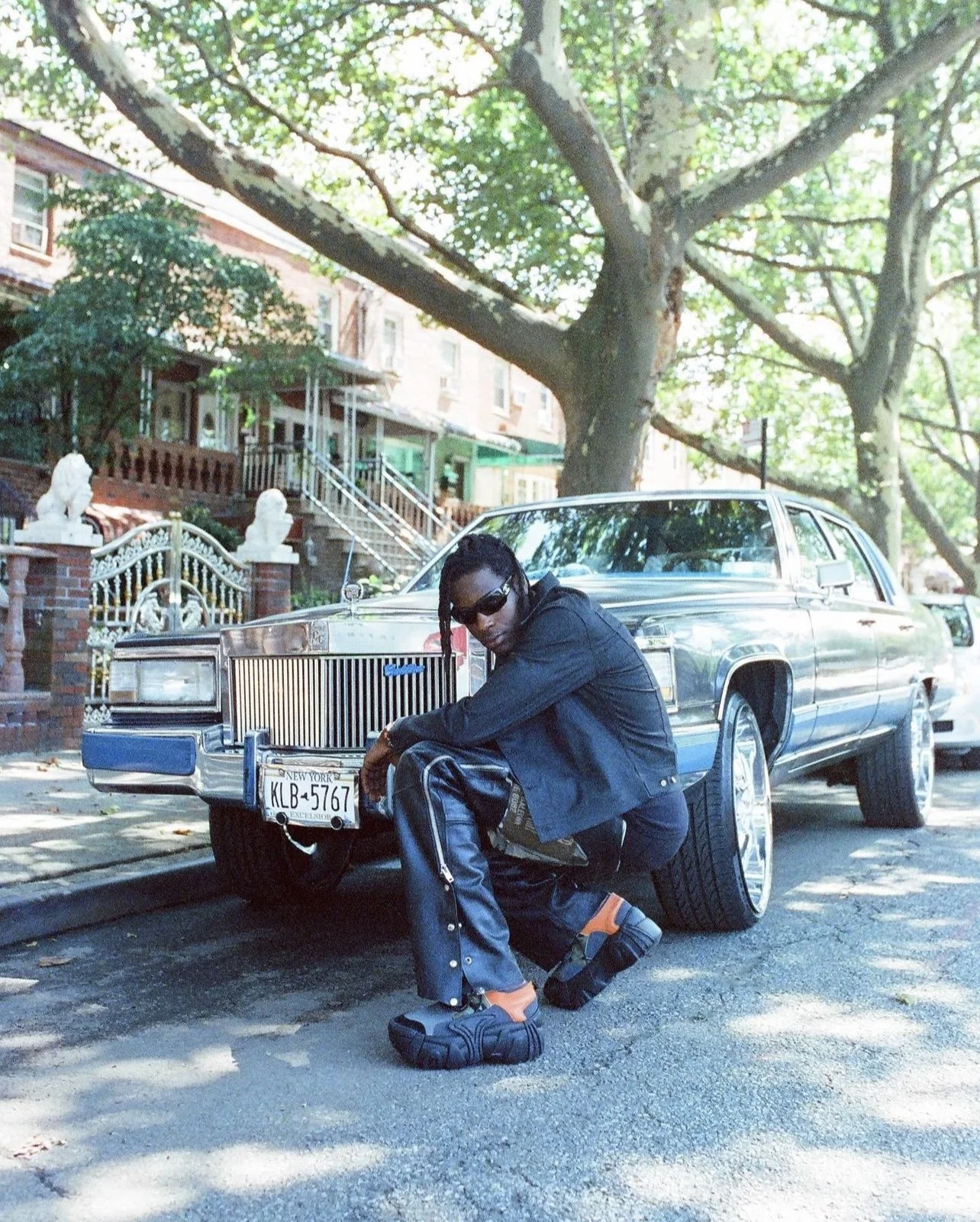 Person wearing sunglasses, black jacket, and baggy pants kneeling in front of a vintage Cadillac parked on a shaded street in a residential neighborhood with trees and row houses.