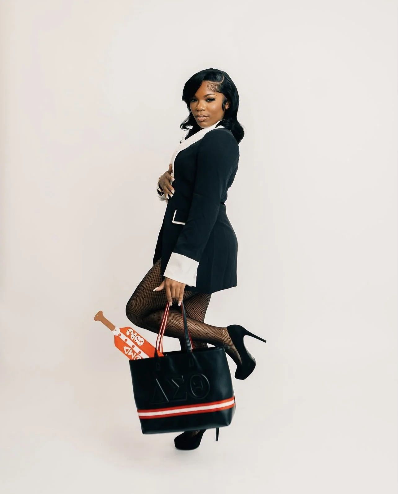 A woman in a black blazer, fishnet stockings, and high heels, holding a black handbag, with her foot resting on a pizza box, standing against a plain white background.