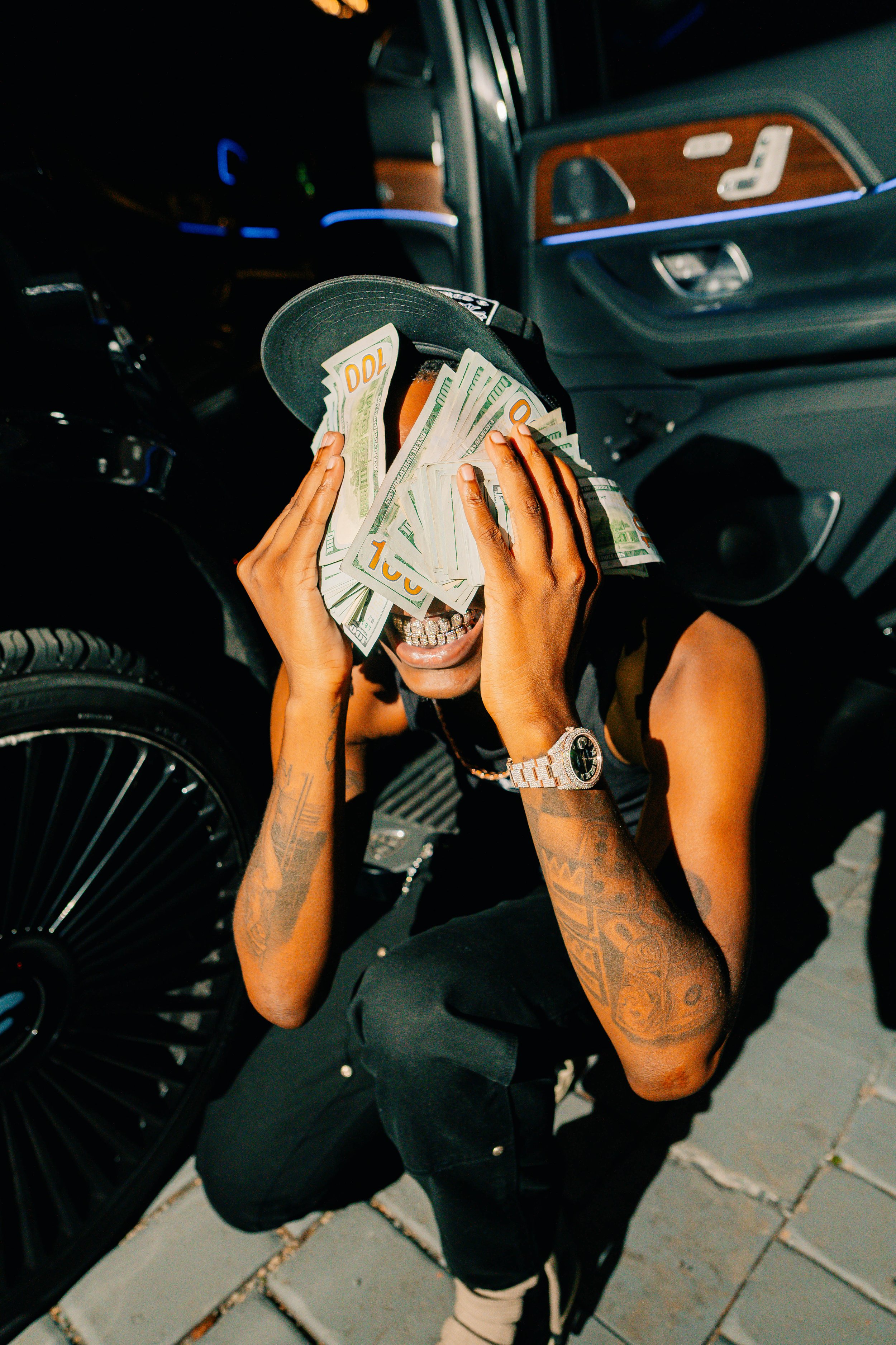 Person kneeling on the ground holding a cap filled with cash over their face inside a car.