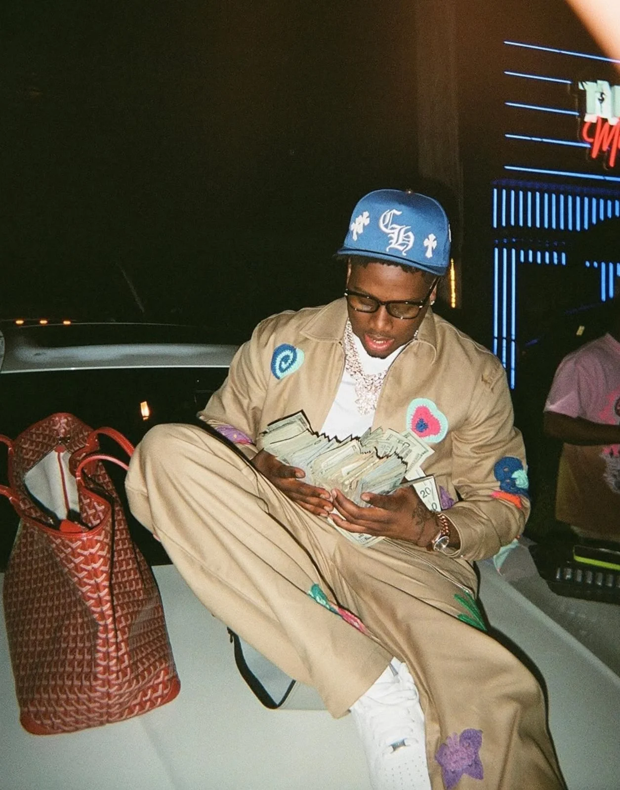 A young man wearing a blue baseball cap, glasses, and designer clothing sits on a ledge at night, holding a large amount of cash. There is a large brown patterned bag next to him and a neon-lit sign in the background.