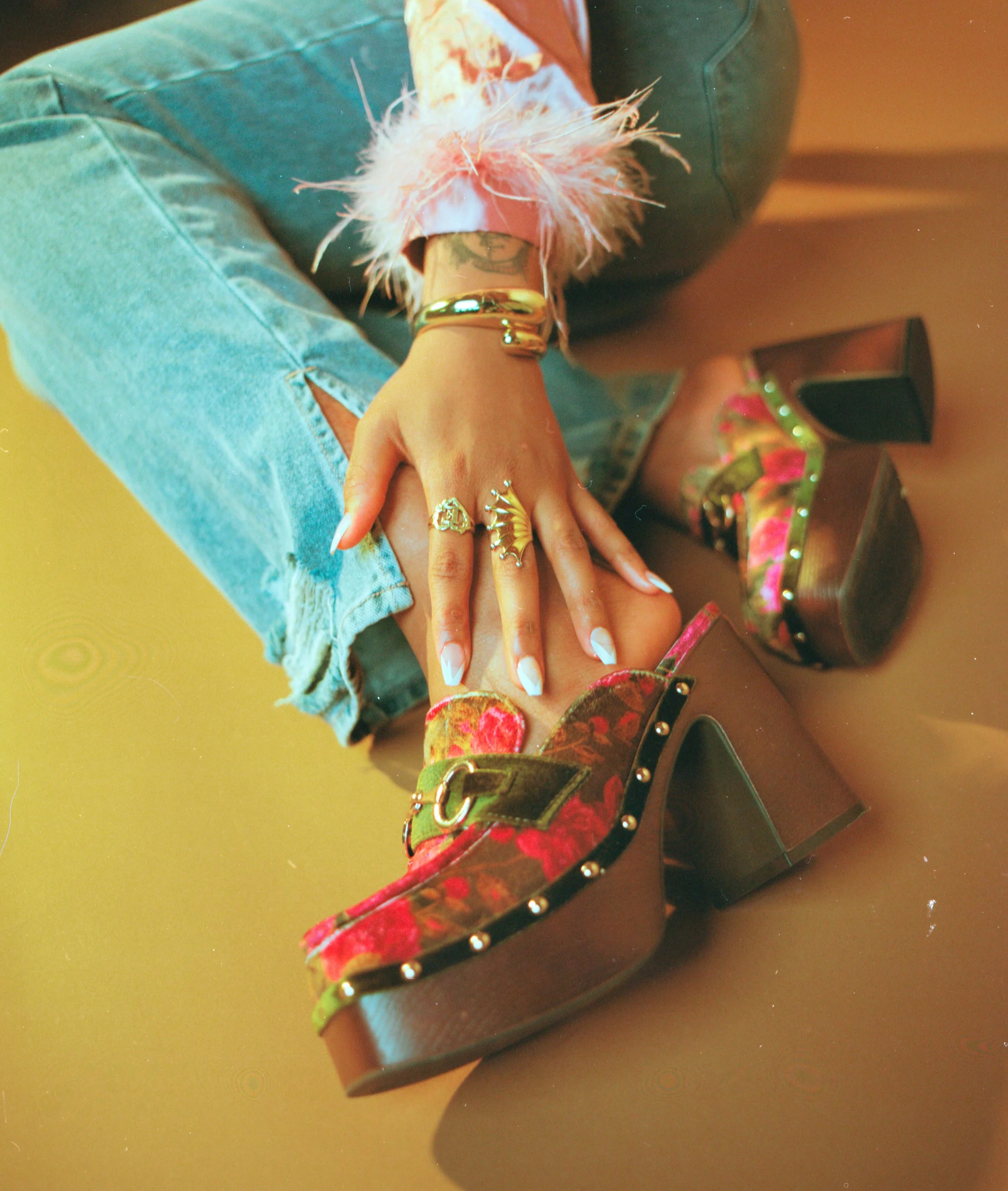 Person wearing floral platform heels with studs, ripped denim jeans, and gold rings, sitting on the floor.