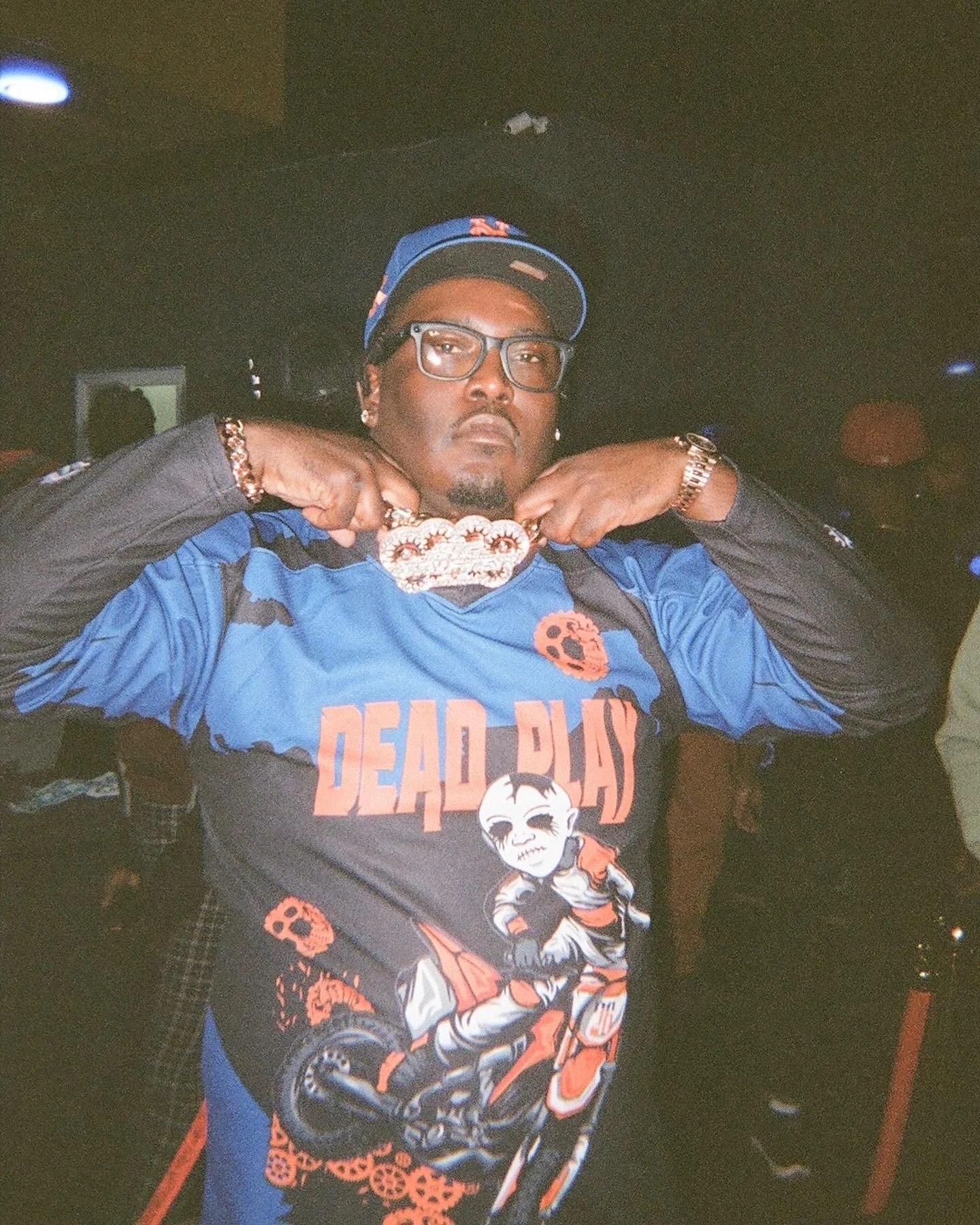 Man wearing a blue and black sports jersey with a graphic design, holding a gold chain with a large pendant, wearing glasses, a blue cap, and jewelry, posing in a dimly lit environment.