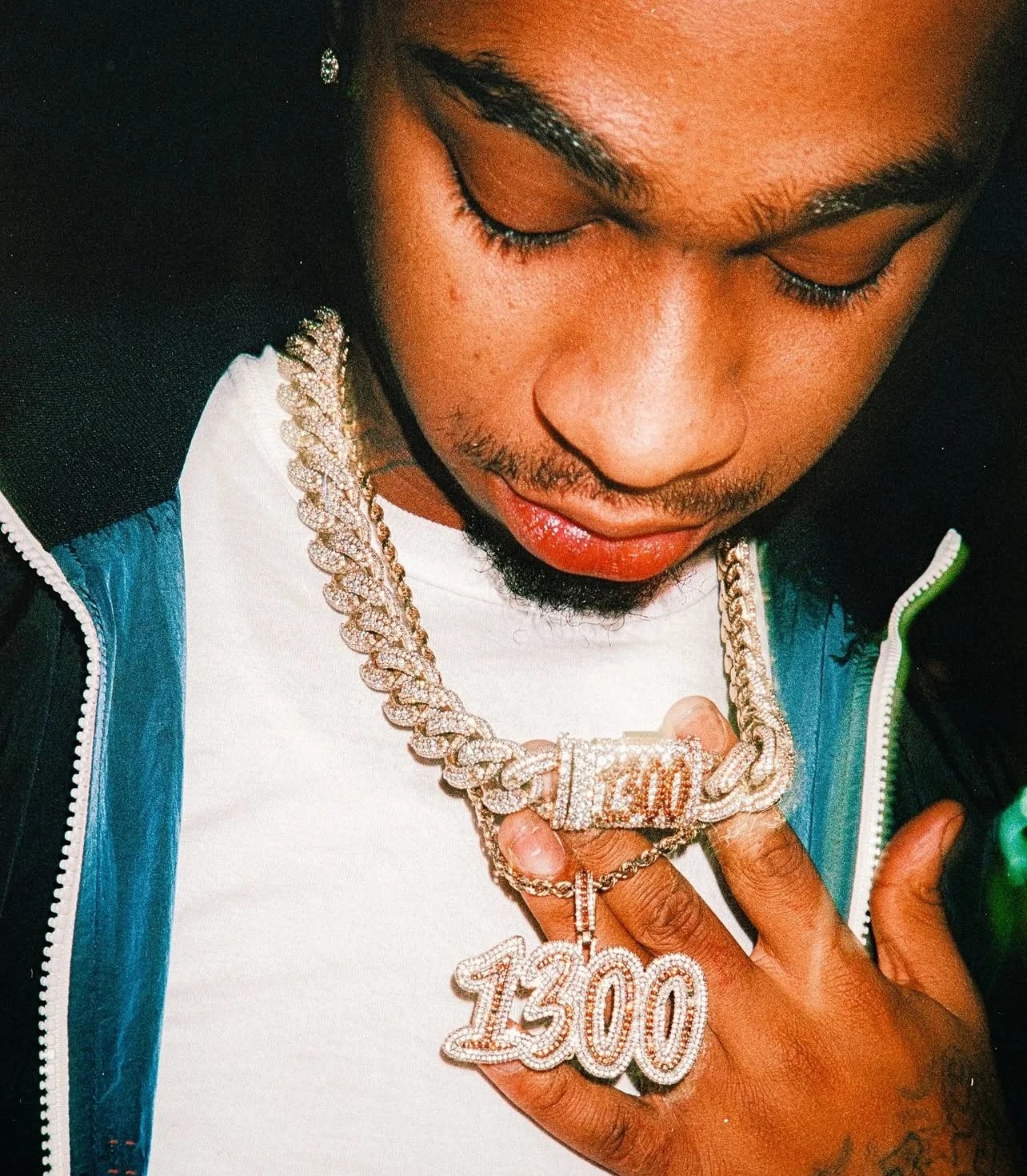 A young man wearing jewelry, including a large chain with a "1300" pendant and a diamond-encrusted clasp, is looking down at his chest.