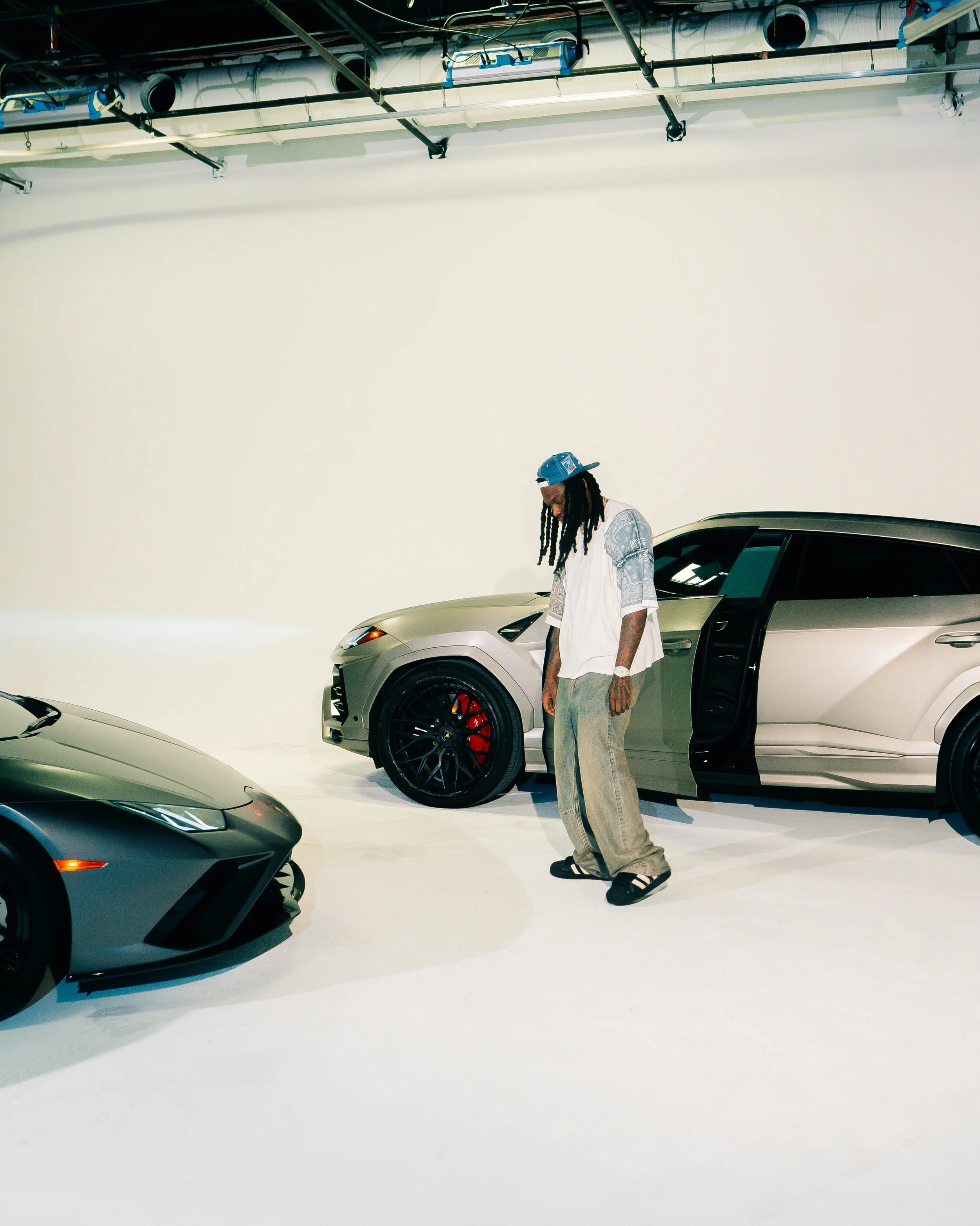 A man in a cap, t-shirt, baggy pants, and sandals stands beside two luxury cars in a studio with a white background and industrial ceiling pipes.