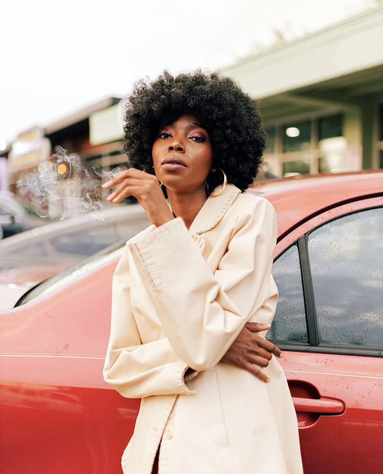 A woman with curly black hair, hoop earrings, and makeup, standing in front of a red car while smoking a cigarette on a rainy day.