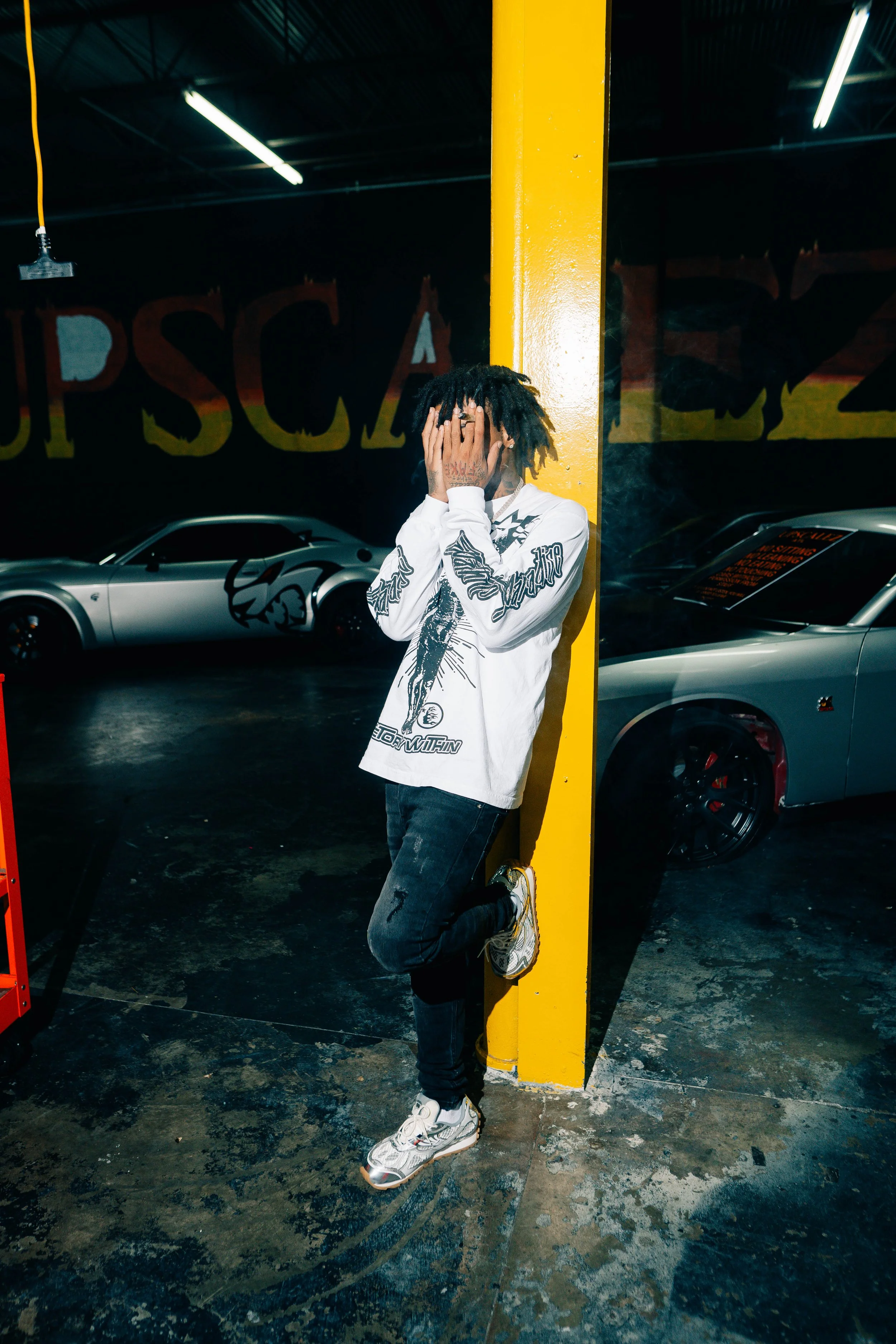 Person with dreadlocks covering face, wearing a white graphic long sleeve shirt, black jeans, and sneakers, leaning against a yellow pillar in a dimly lit garage with sports cars in the background.