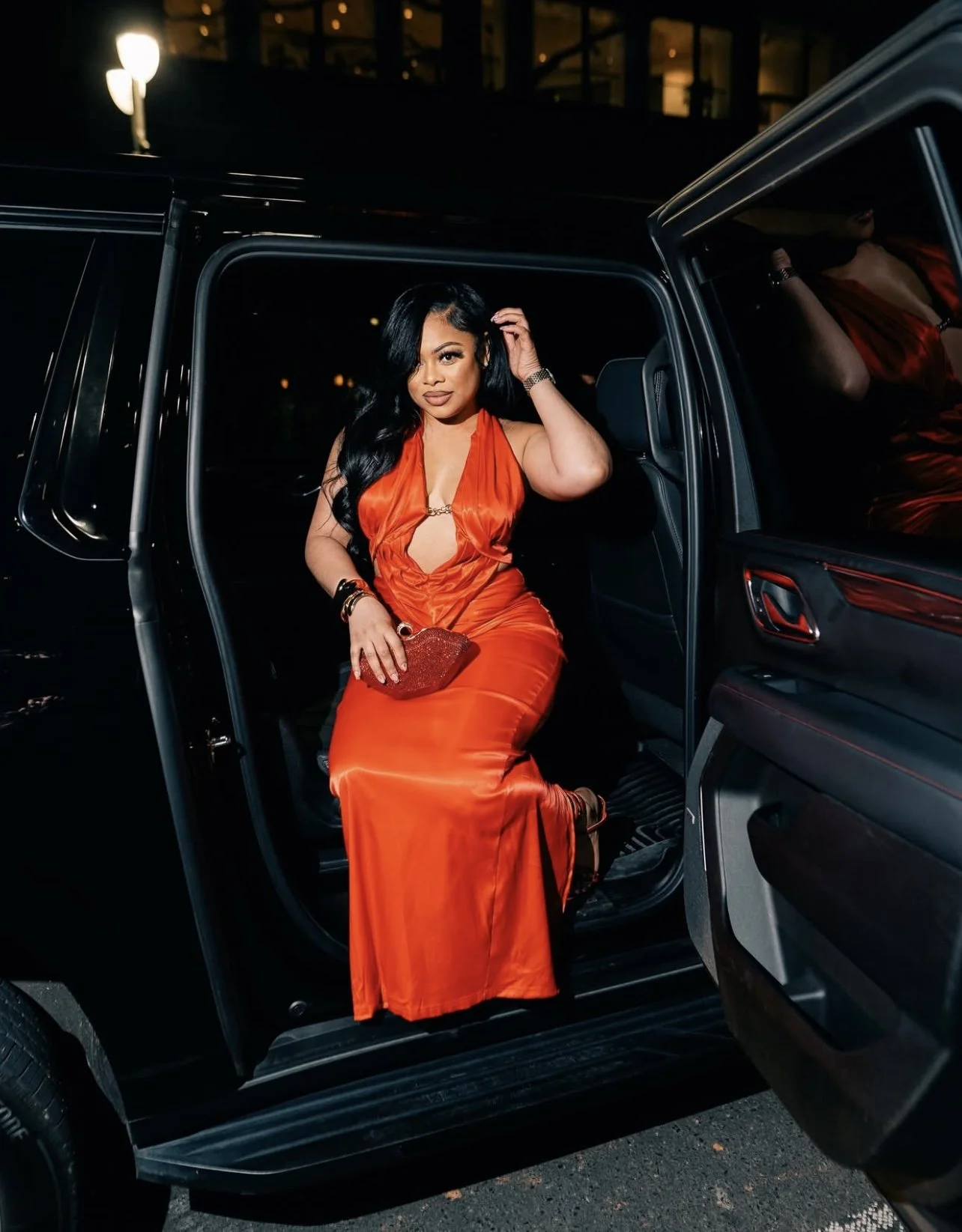 A woman in an orange satin evening gown sitting in the open door of a black vehicle at night, holding a red clutch purse, with city lights and a building in the background.