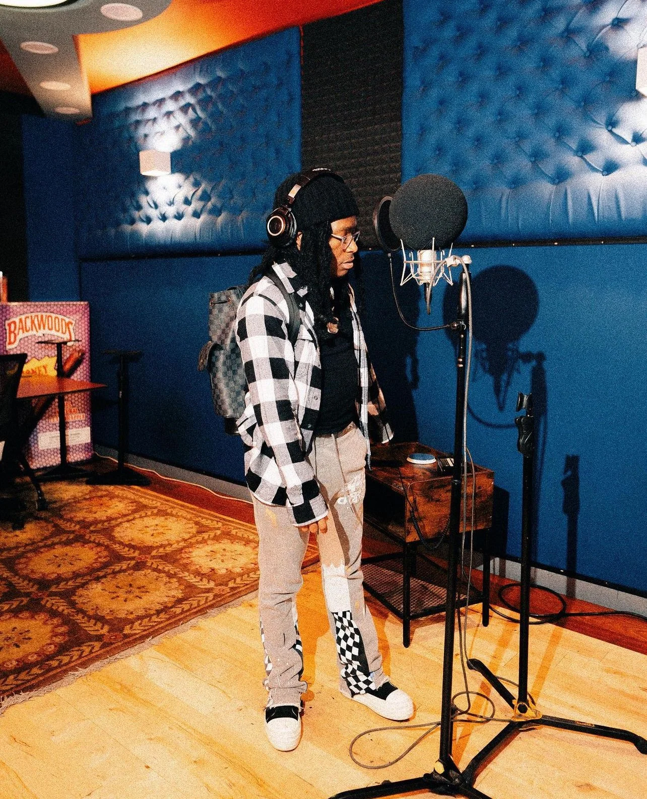 A person with long dreadlocks, wearing glasses, headphones, a black beanie, a black and white checkered jacket, beige pants with checkered patches, and white sneakers, stands in a music recording studio in front of a microphone.