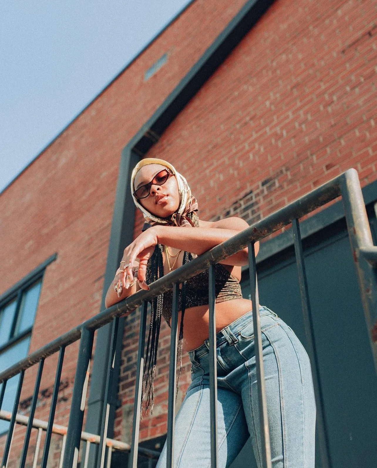 A young woman wearing sunglasses, a headscarf, a black crop top, and jeans leaning on a metal railing outside a brick building.