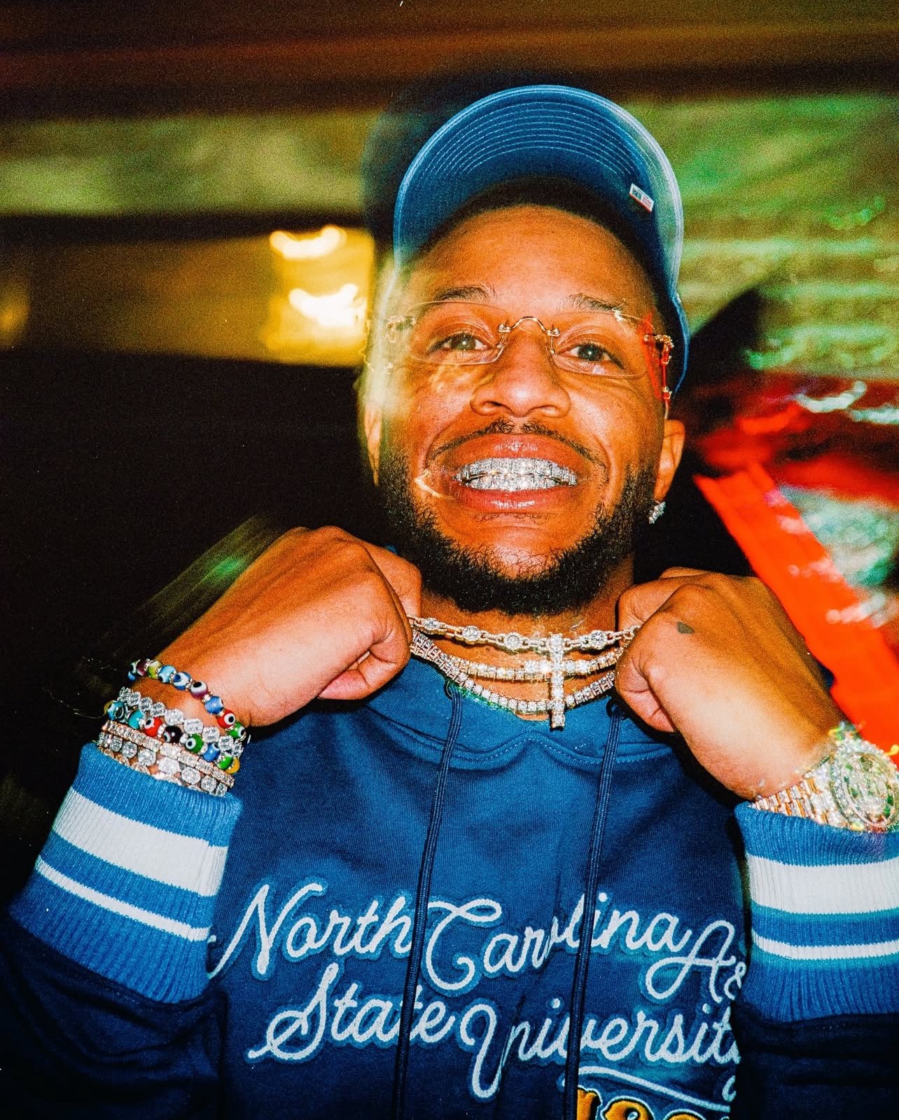 A smiling man wearing a blue North Carolina State University hoodie, a blue cap, and multiple jewelry pieces including bracelets, chains, and a watch, holding his necklaces with both hands.