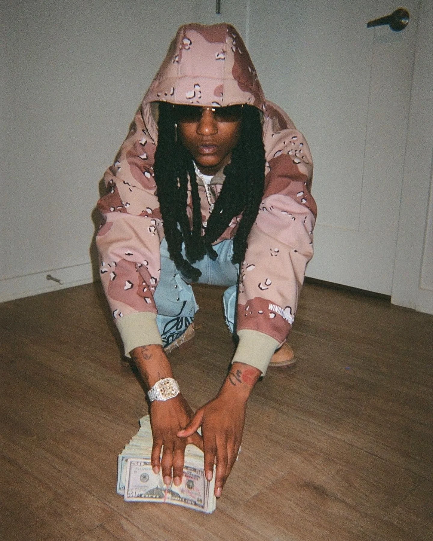 A person with long braids crouching on the floor, wearing sunglasses, a camouflage hoodie, and jeans, holding a stack of money.