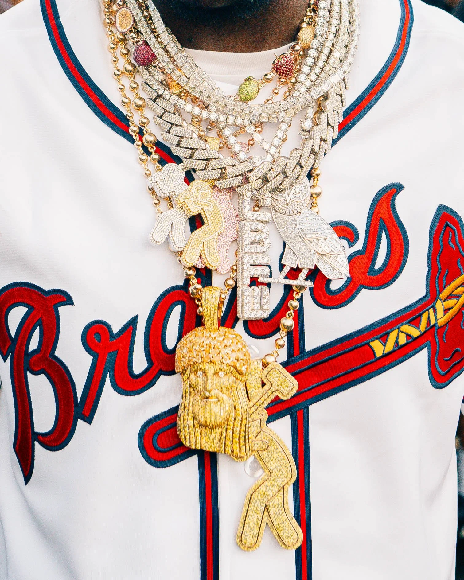 Close-up of a person's chest wearing multiple gold and diamond-encrusted necklaces and chains, with a baseball jersey featuring red and blue accents and red script reading 'Braves'.