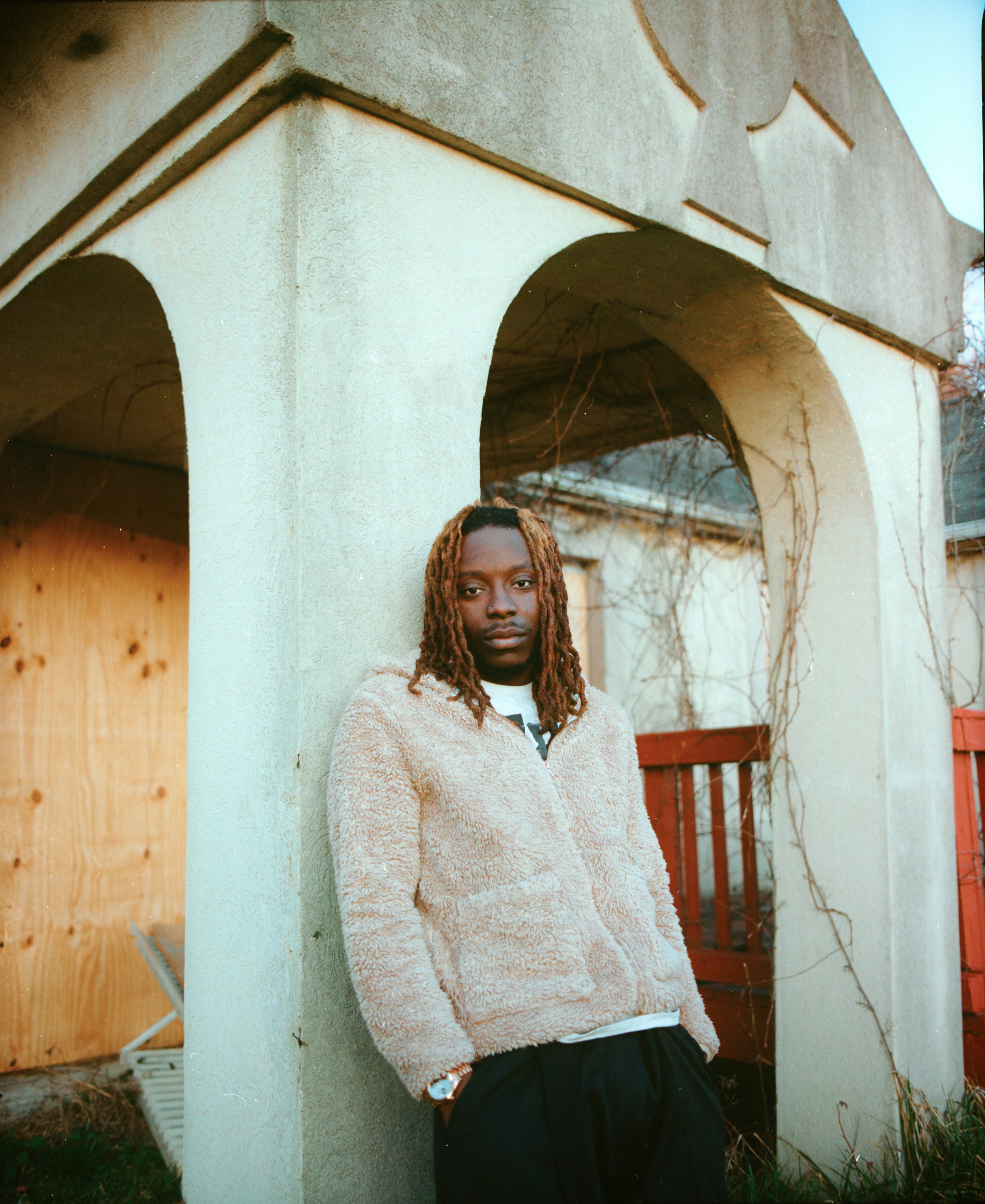 A person with dreadlocks wearing a beige fuzzy jacket and black pants standing and leaning against a corner of a stucco wall with arched openings, with a wooden fence and grapevines in the background.