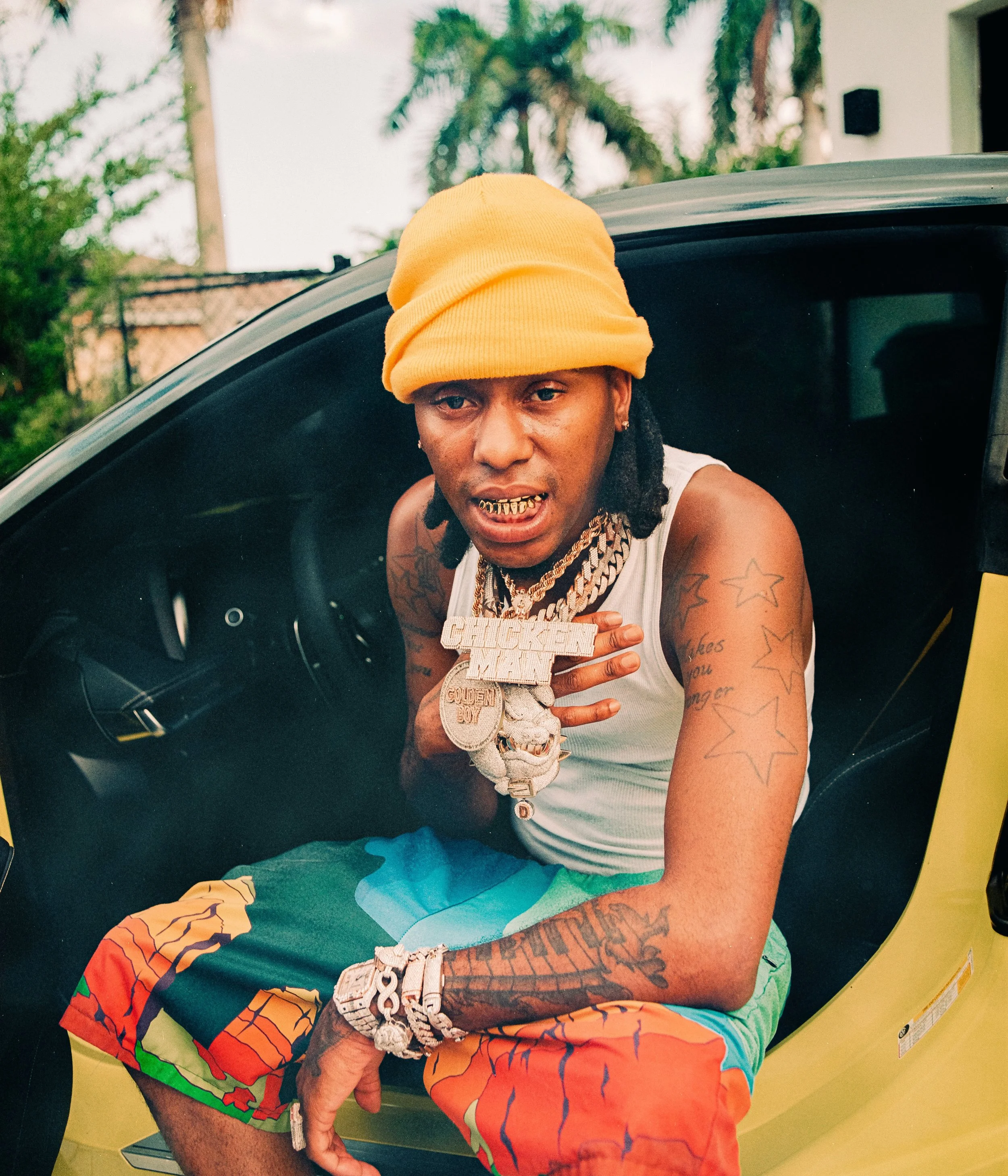 A man with tattoos, gold chains, and jewelry wears a yellow beanie and colorful shorts while sitting in a black and yellow sports car. He is making a gesture with his hand and looking at the camera.