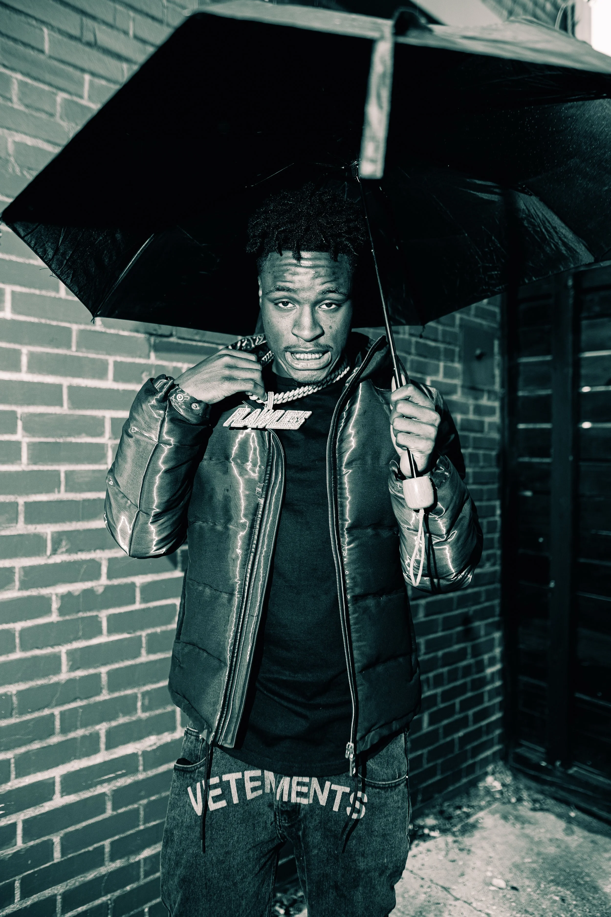 Young man with dreadlocks holding an umbrella, wearing a puffer jacket and jewelry, standing against a brick wall.