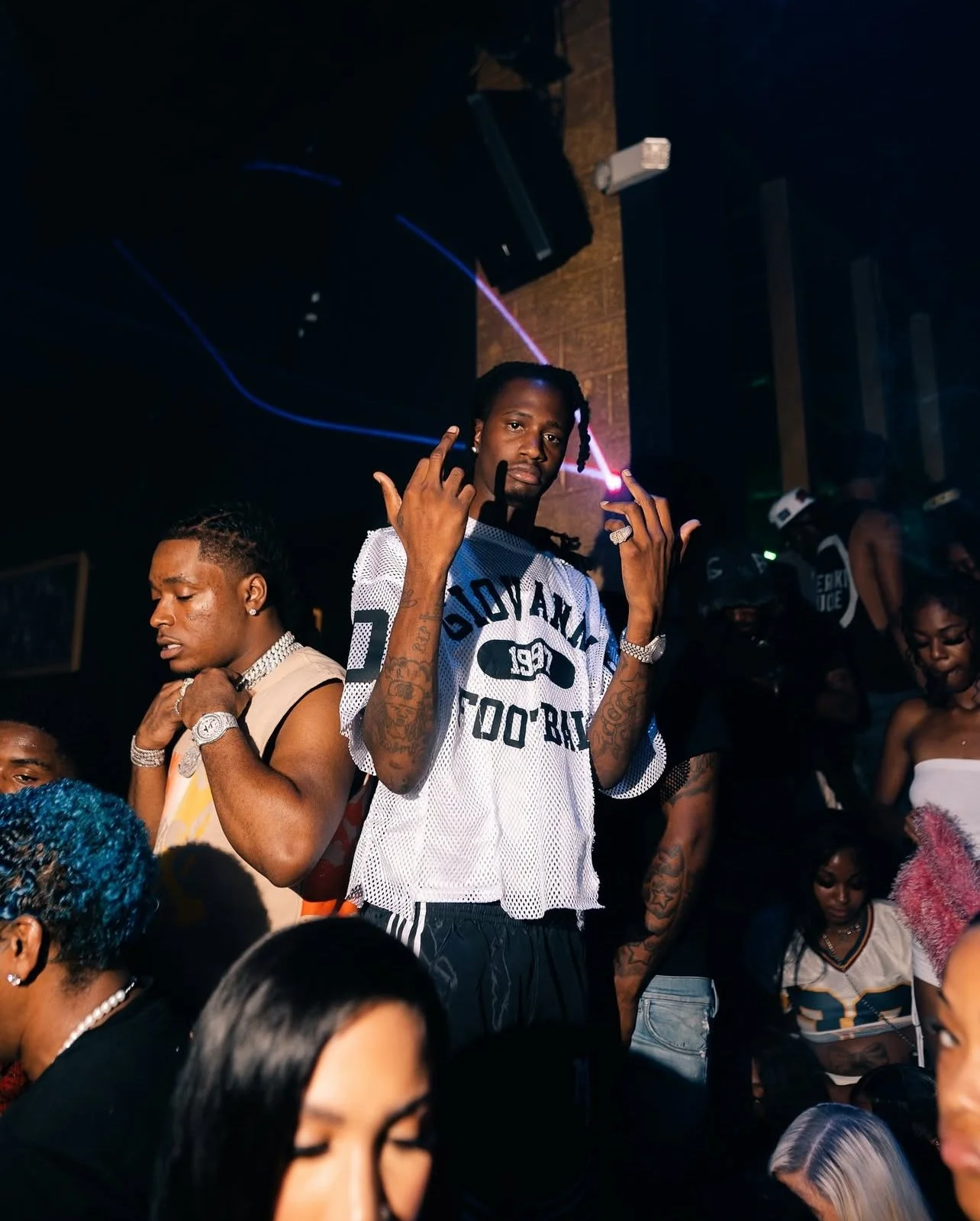 A young man with tattoos making a hand gesture at a nightclub, with other people dancing and socializing around him.