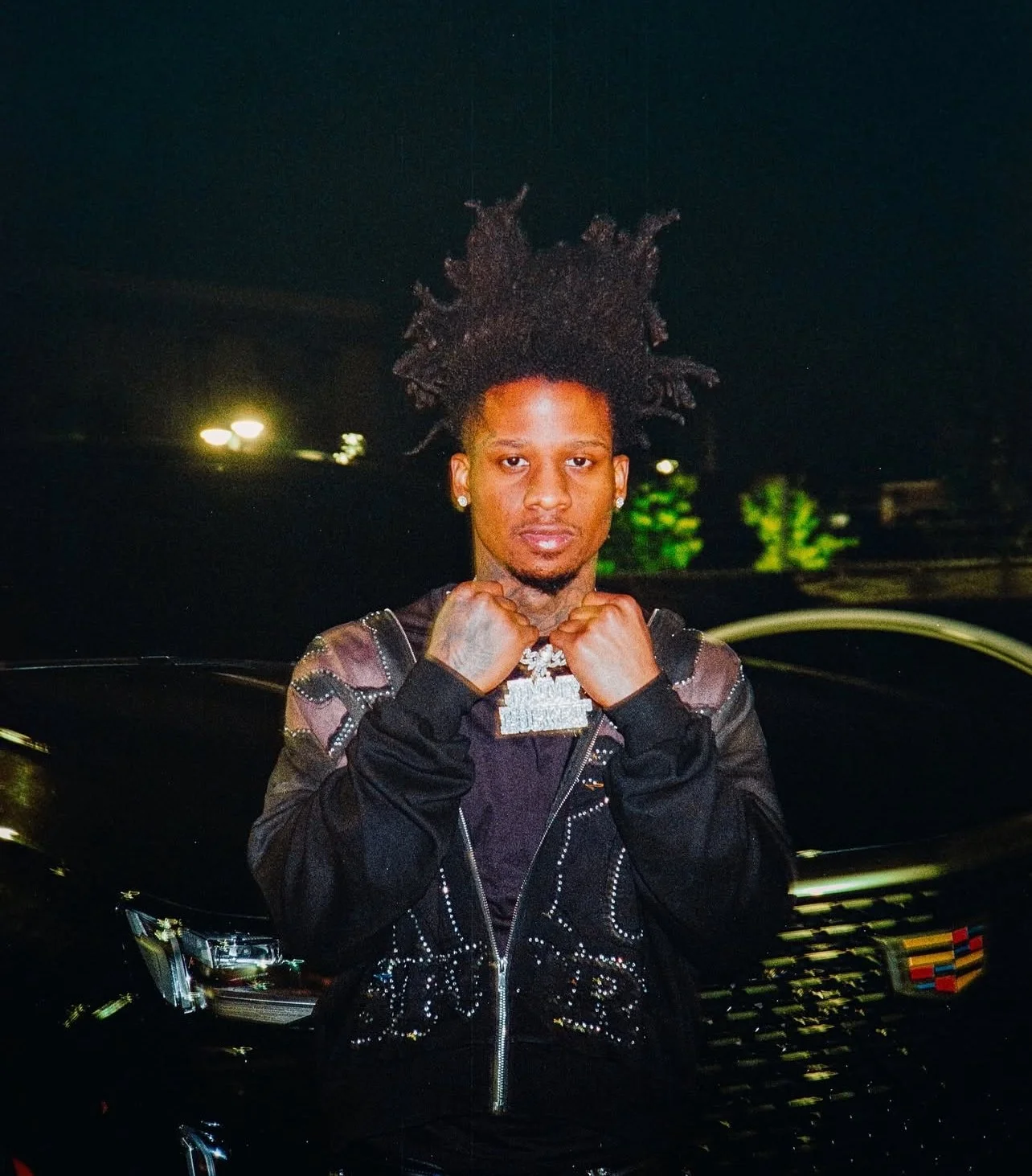 A young man with styled hair, wearing a black jacket with embellishments, standing in front of a dark background with lights and green trees, showing a confident pose.