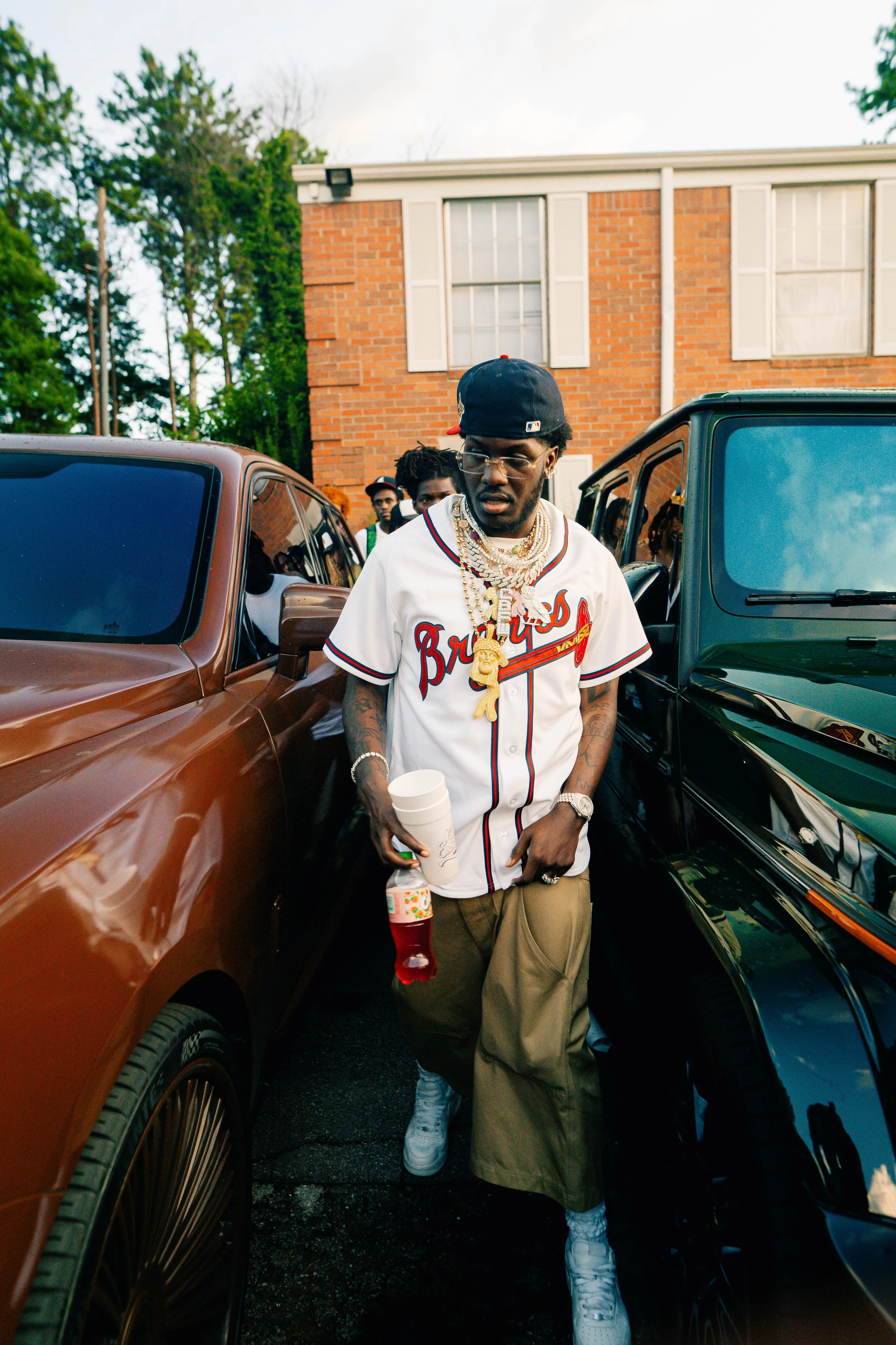 A man in a baseball cap and jewelry standing between two parked cars in front of a brick house with white shutters.