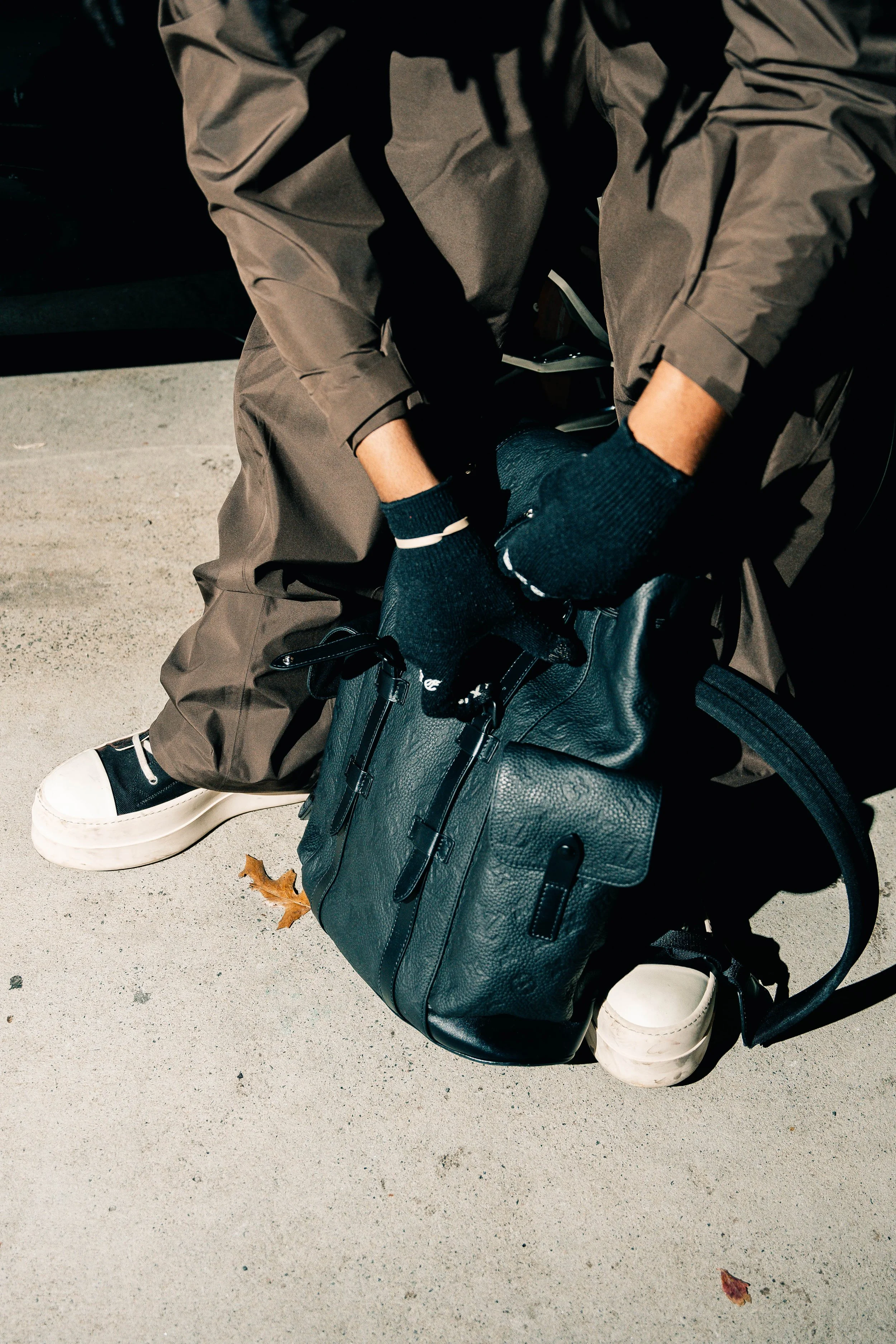 Person sitting on a concrete surface, packing a black leather backpack, wearing gray pants, black gloves, and black and white sneakers, with a dried leaf nearby.