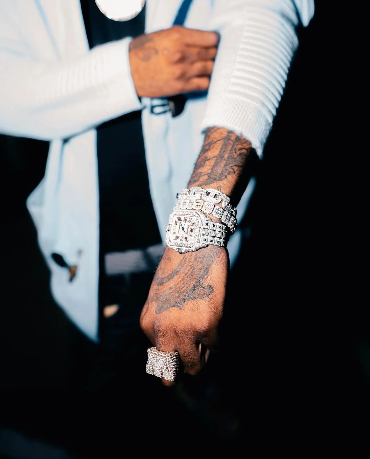 Person displaying a large diamond-encrusted watch on their wrist, wearing a large diamond ring, with tattoos visible on their arm, dressed in a white and black outfit.