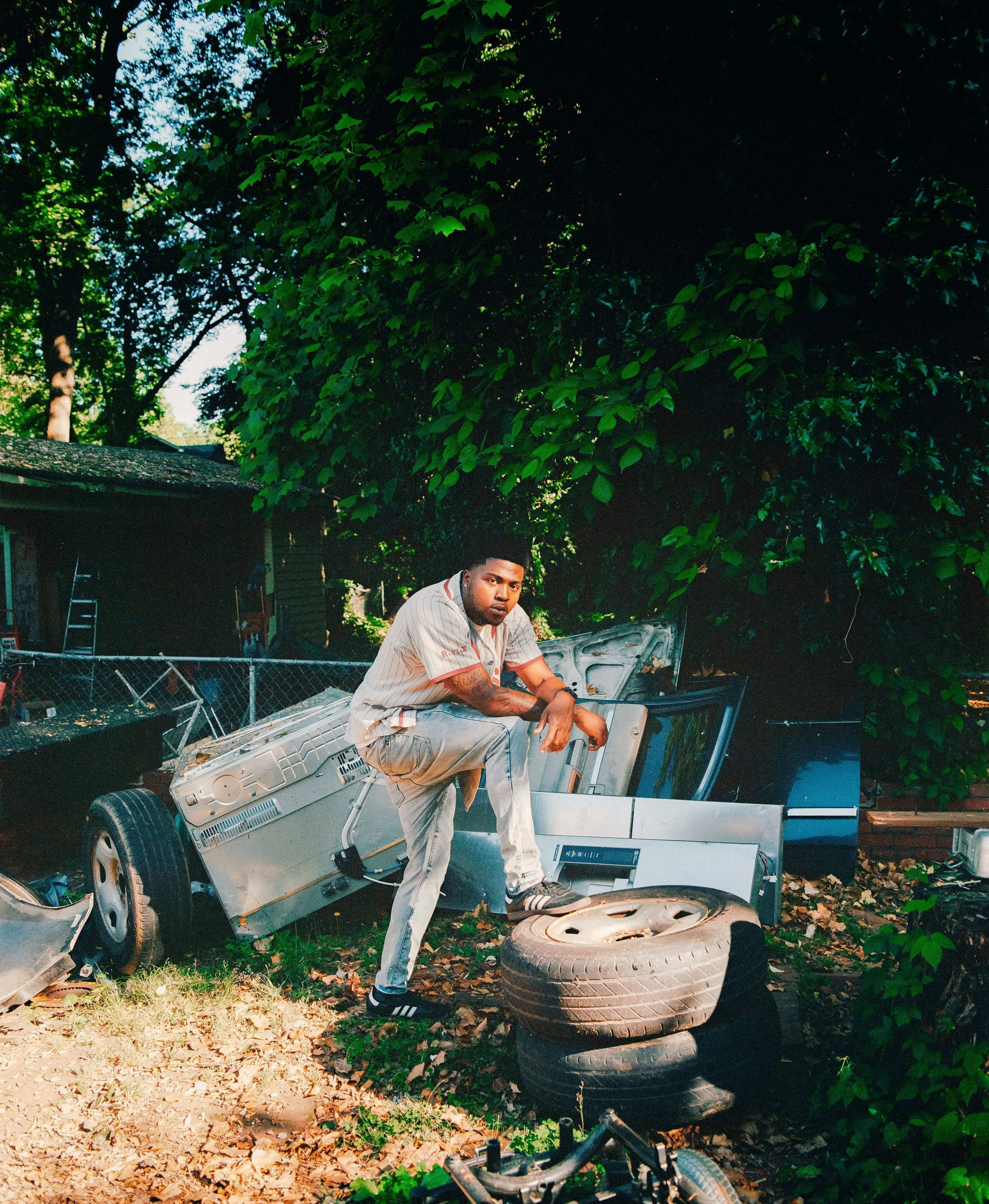 A man with tattoos crouching on a pile of old car parts and tires outdoors next to a large leafy tree.