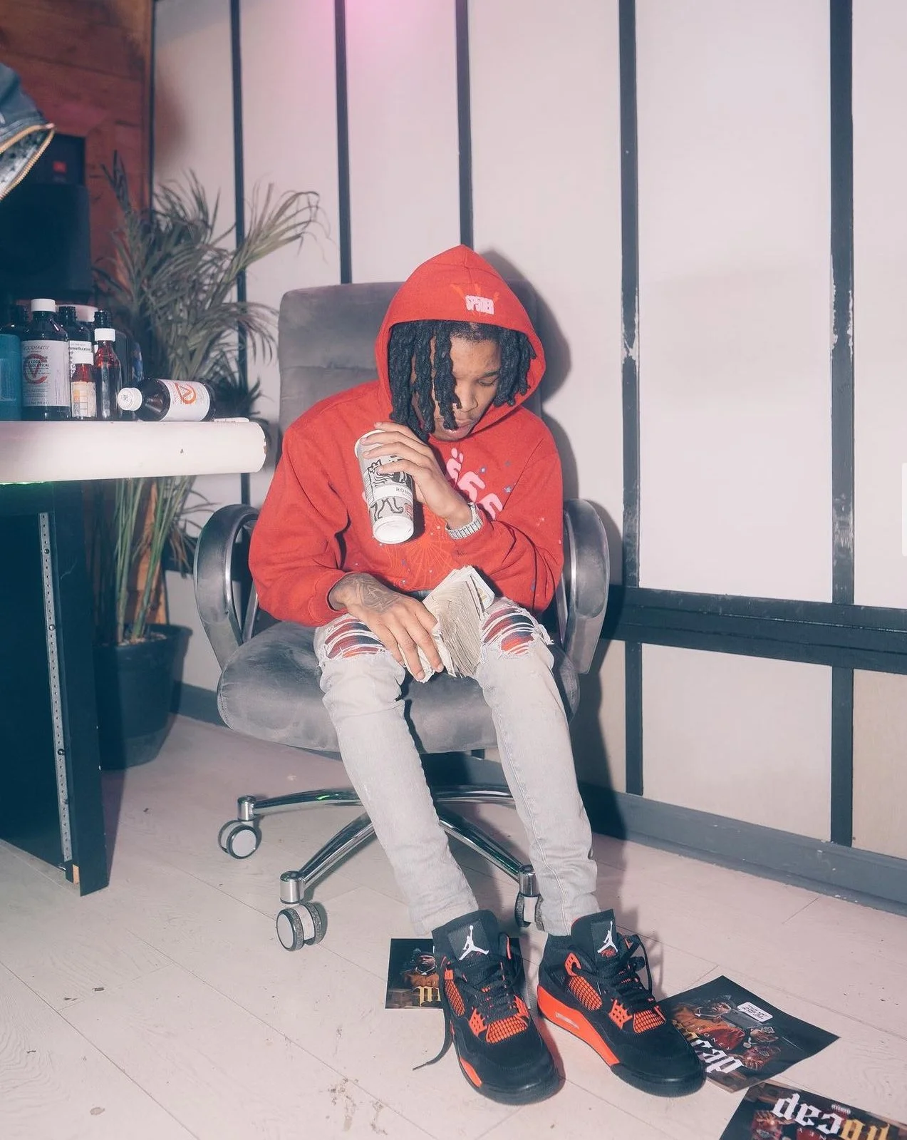 A young man with dreadlocks wearing a red hoodie and ripped light gray jeans, sitting in an office chair, holding a can and a stack of money, with sneakers on his feet, surrounded by magazines on the floor.
