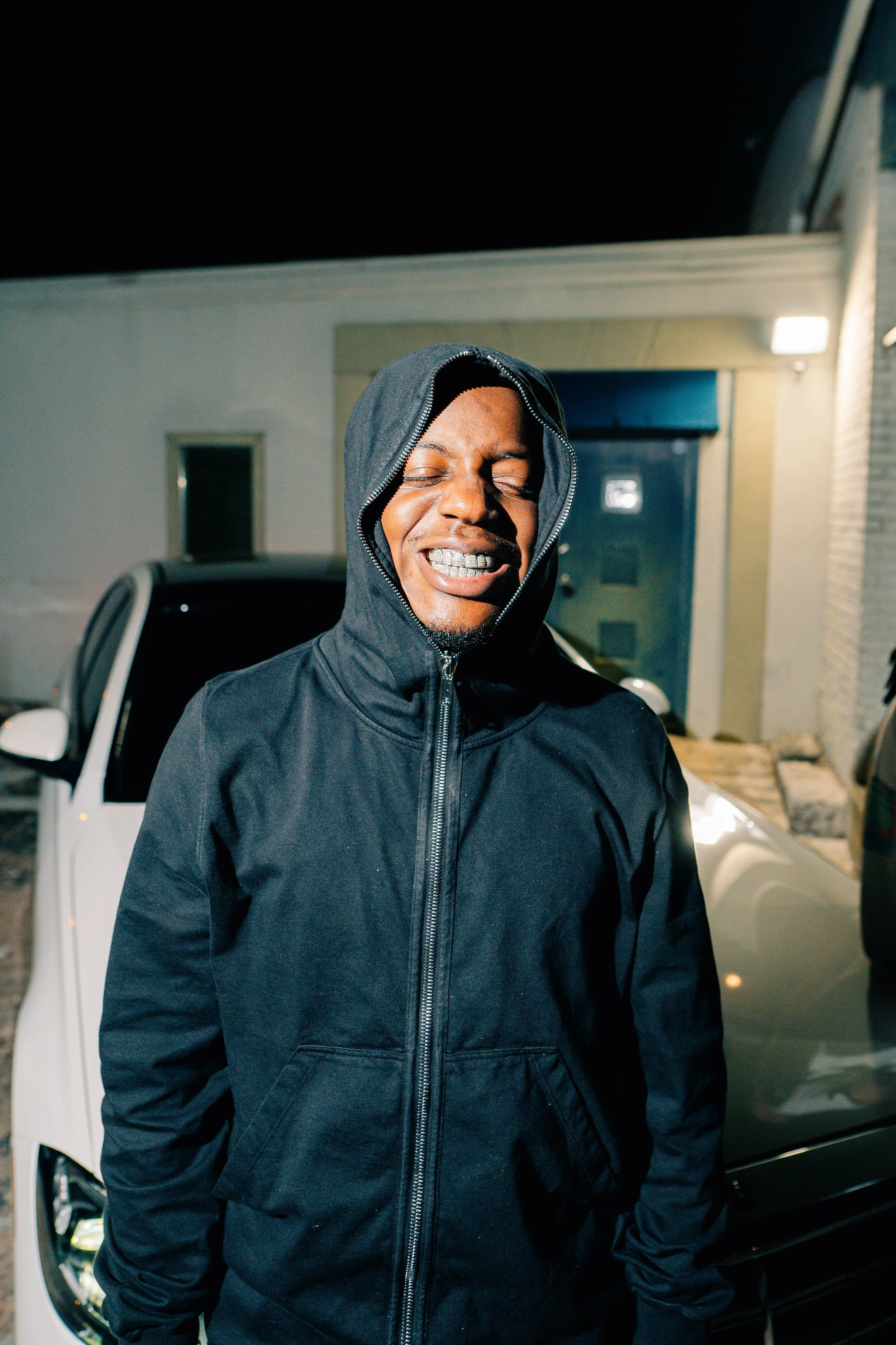 A young man wearing a black hoodie with the zipper partially open, smiling with his eyes closed, standing outdoors at night in front of a white car and a building.