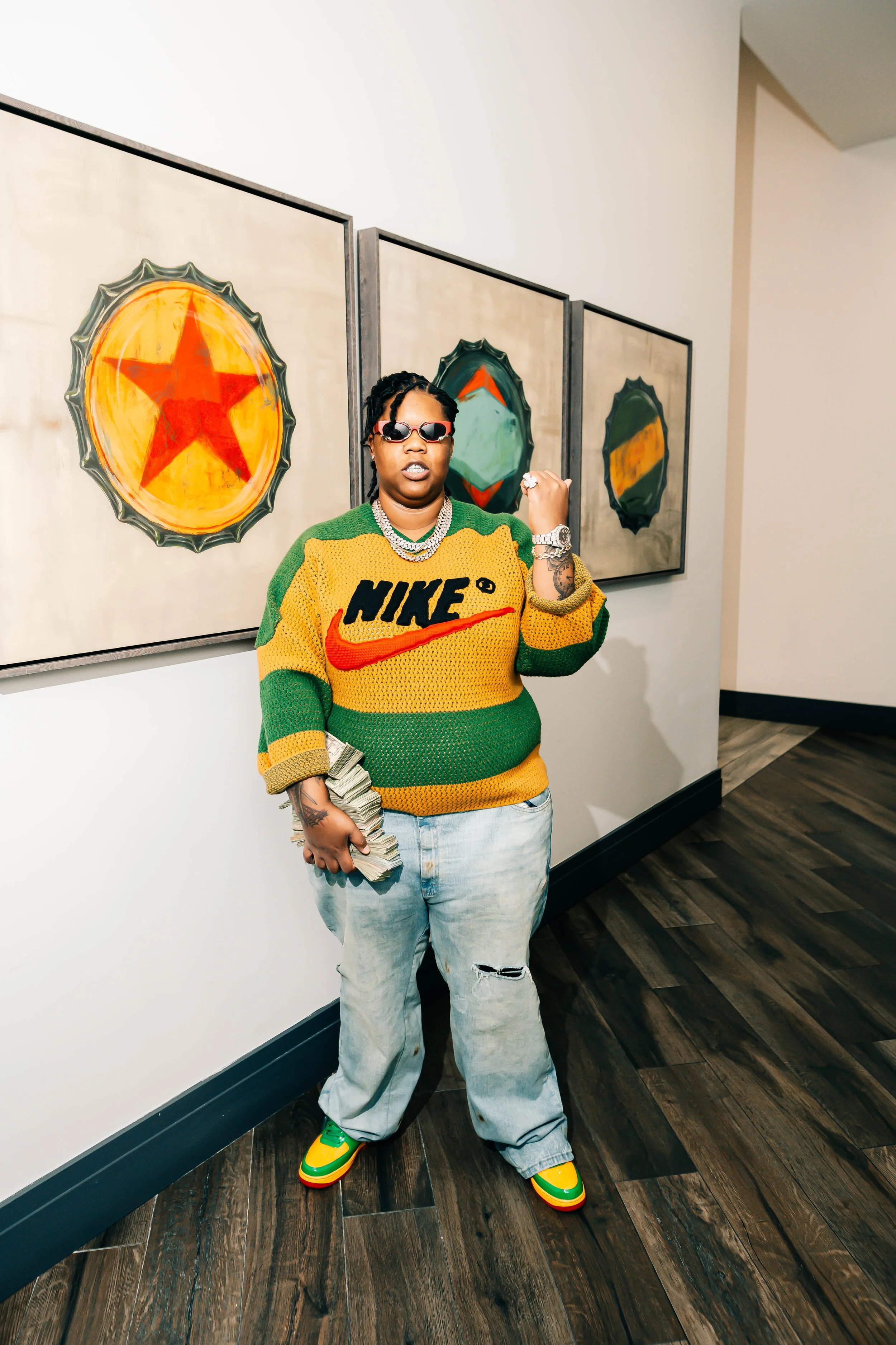 A person standing indoors dressed in a colorful green and yellow sweater, light-washed ripped jeans, and multicolored sneakers, holding a stack of money and wearing sunglasses, with artwork of stars and shapes on the wall behind them.
