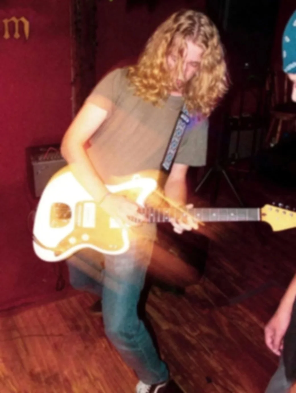 A person with long curly hair playing an electric guitar in a dimly lit room with wooden flooring.