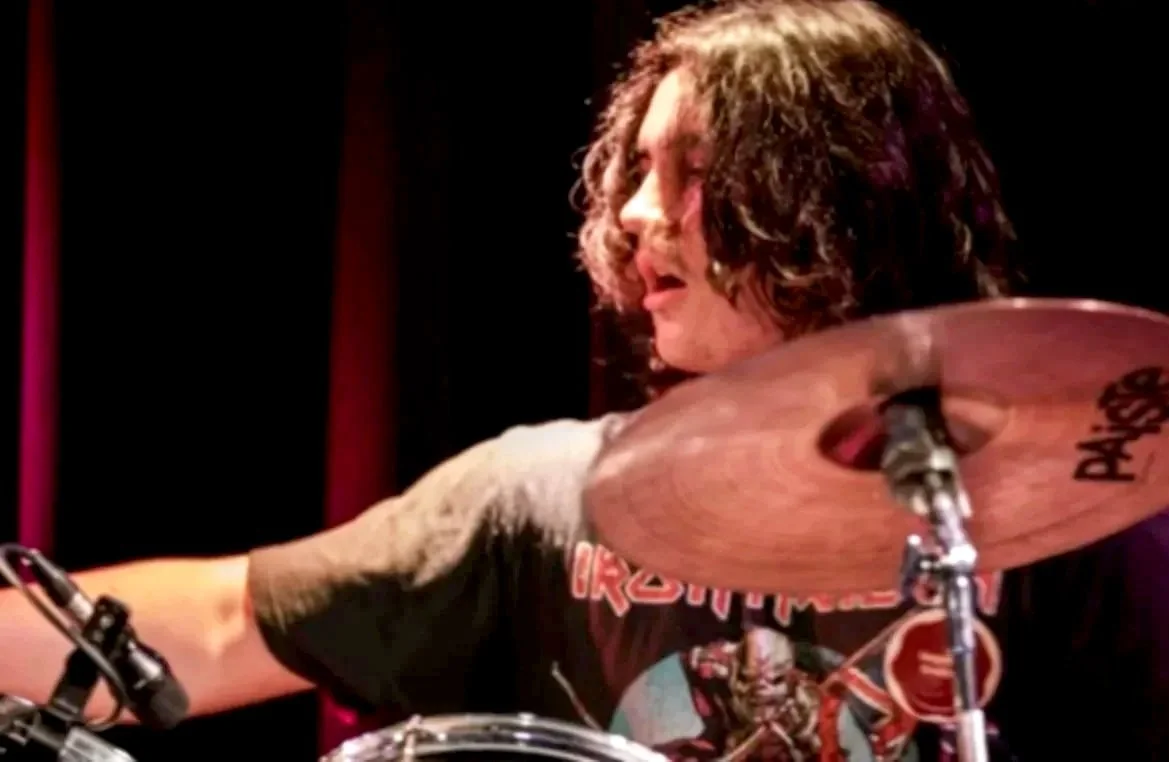 A man playing drums with curly hair and wearing a black band T-shirt, focused on playing the cymbal in a dark setting.