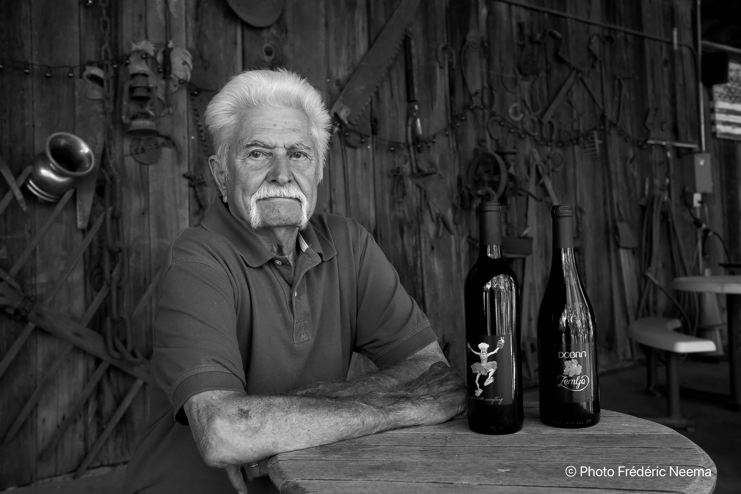 Owner and winemaker Milan sits at a round wooden table in front of a barn with tools hanging on a wooden wall behind him. On the table are two wine bottles.