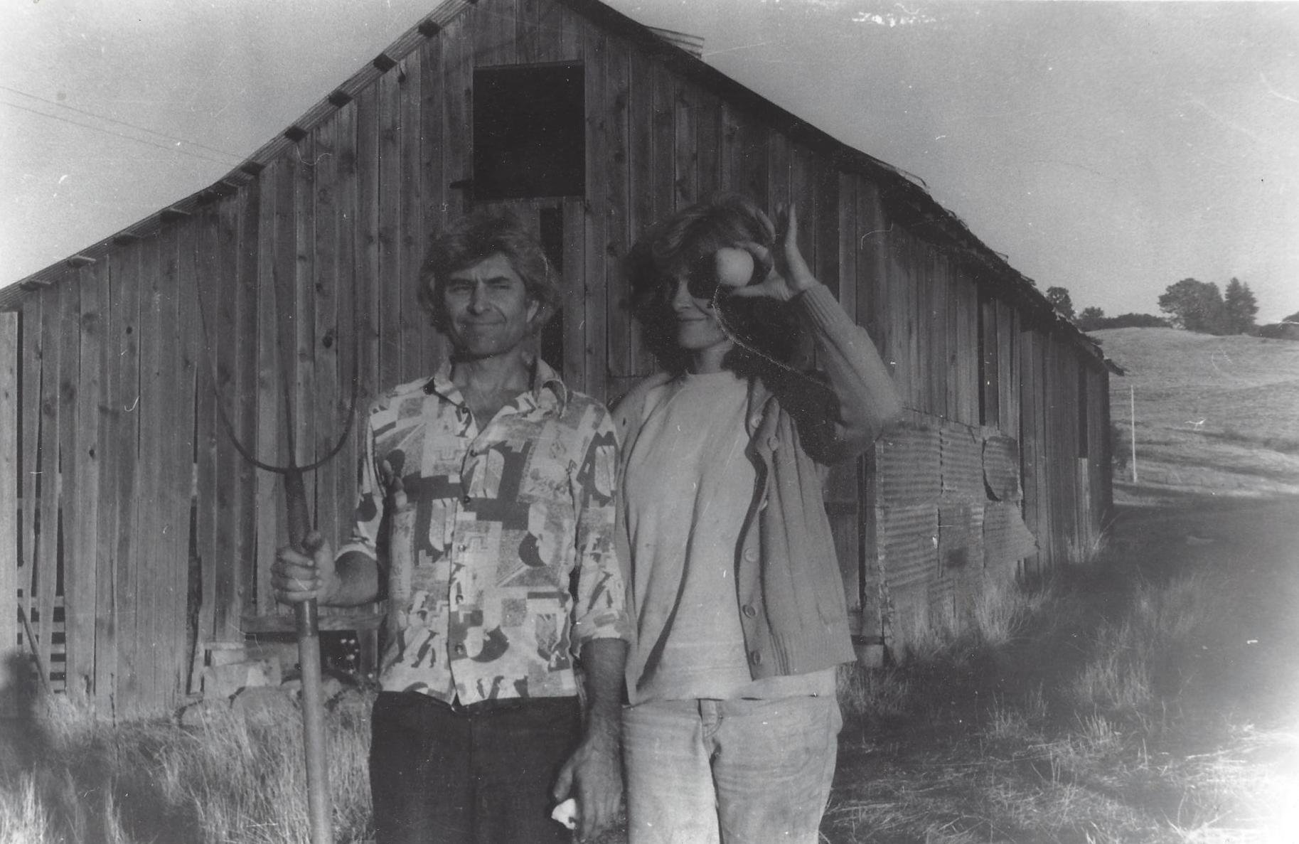 A man and woman standing outdoors in front of a wooden barn, the man holding a pitchfork the woman holding an apple towards her eye, both smiling in black and white photography.